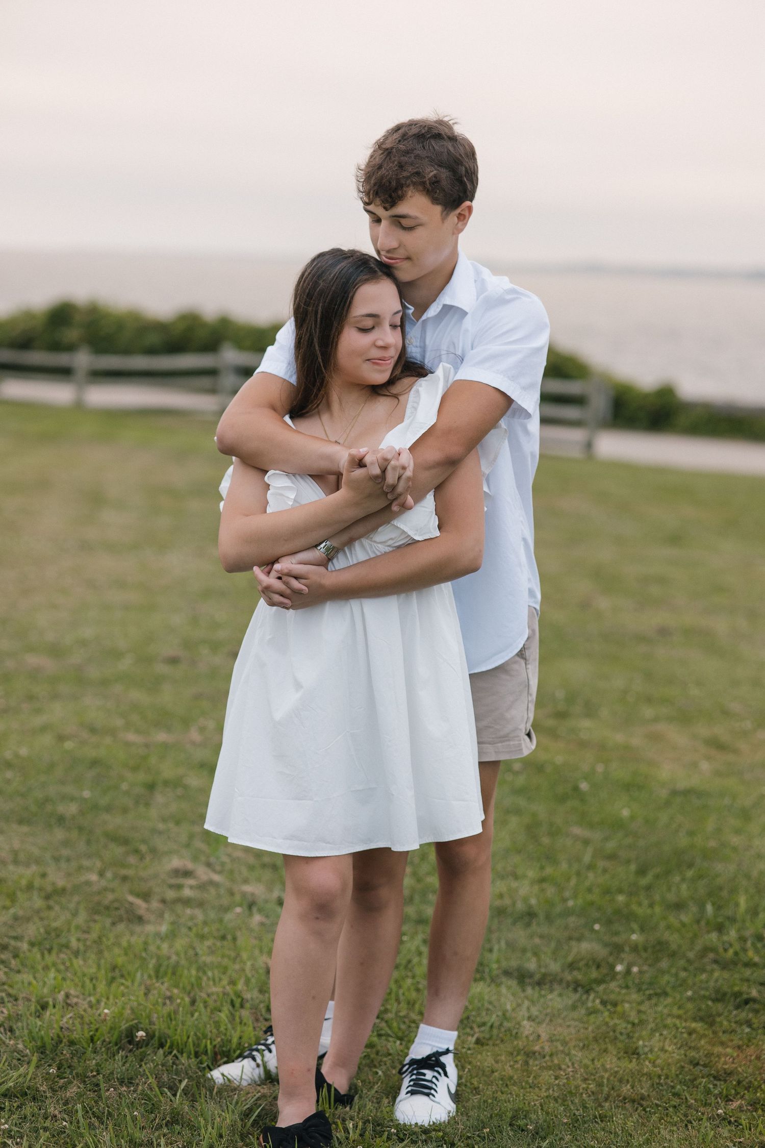 A romantic couple embraces on a scenic coastal overlook while wearing matching white summer outfits.