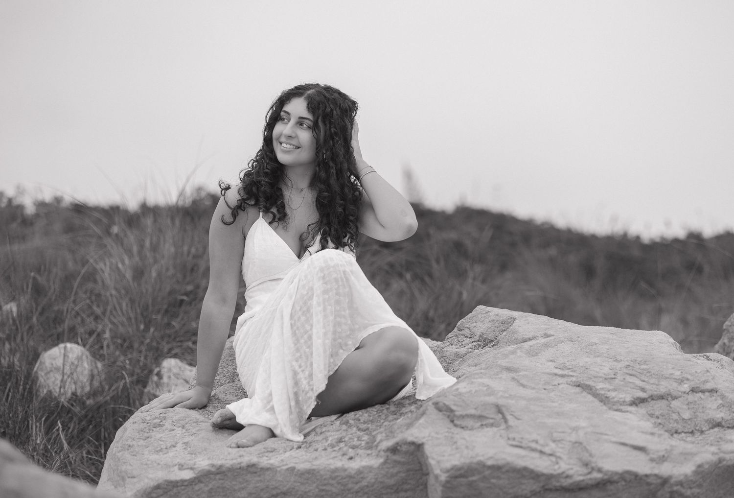 A woman in a white dress lounges gracefully on a rocky beach in a series of artistic black and white photographs.