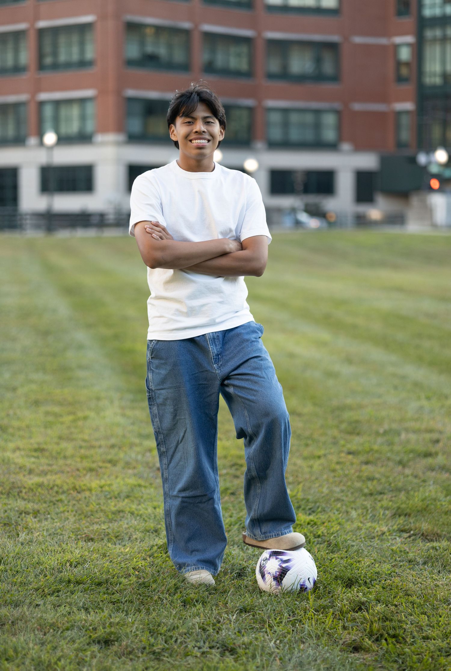 Person poses with soccer ball on grass field wearing white t-shirt and jeans.