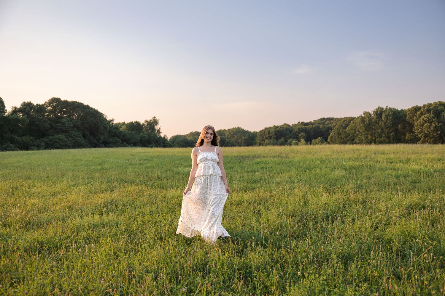 Woman in white dress standing in a lush green field at sunset with trees in the background.