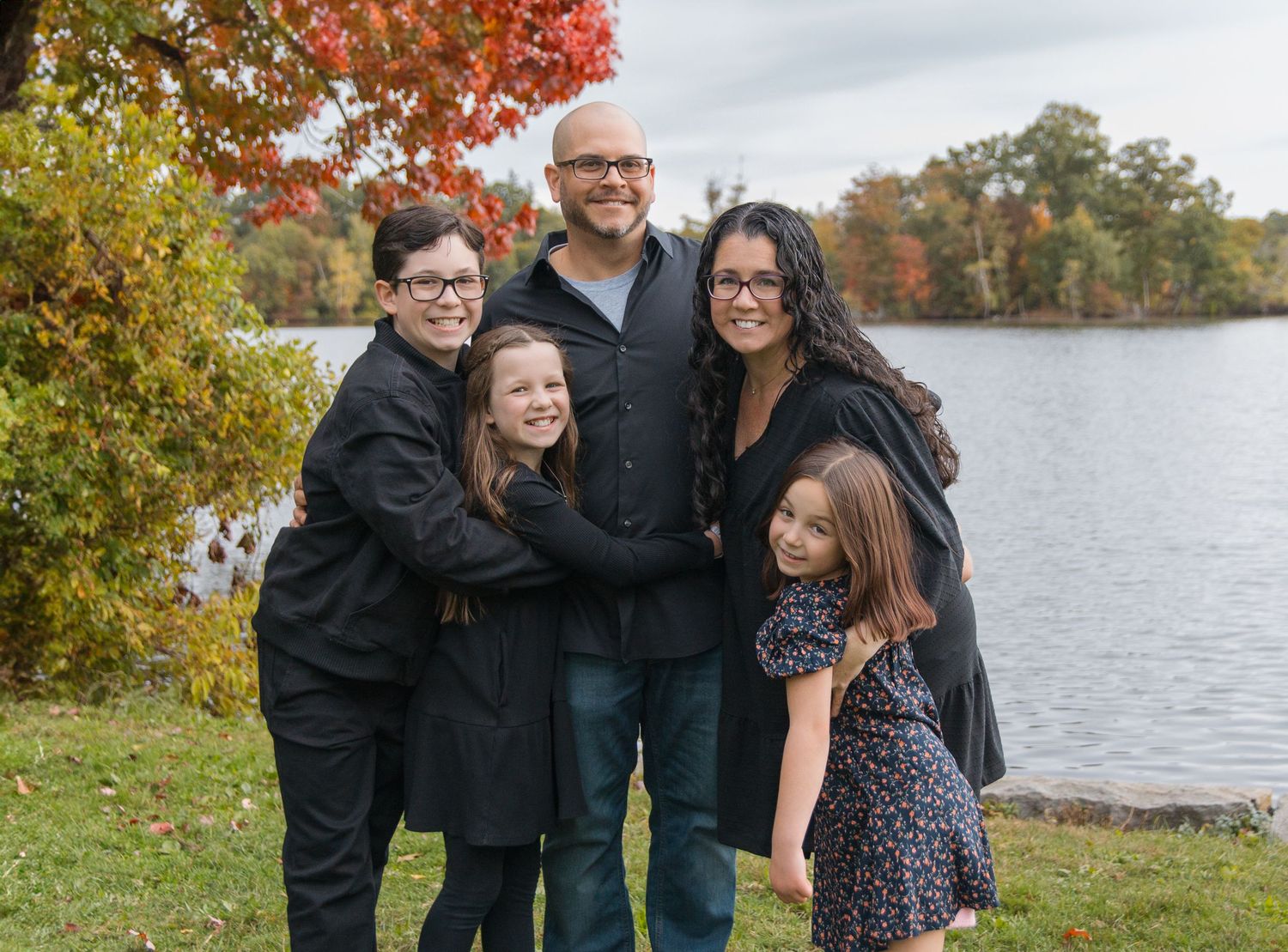 A family group poses together by a lake with autumn colors in the trees.
