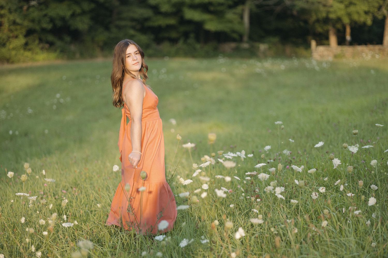 A person in a flowing coral dress stands in a sunny meadow filled with white wildflowers and tall grass.