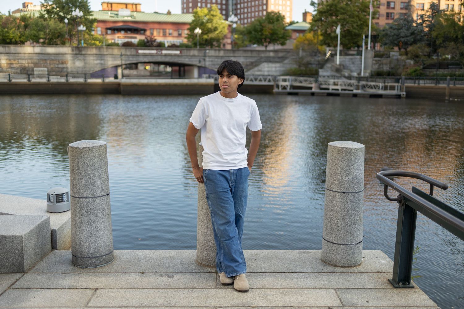 Person stands between stone pillars at waterfront wearing white t-shirt and jeans at sunset.