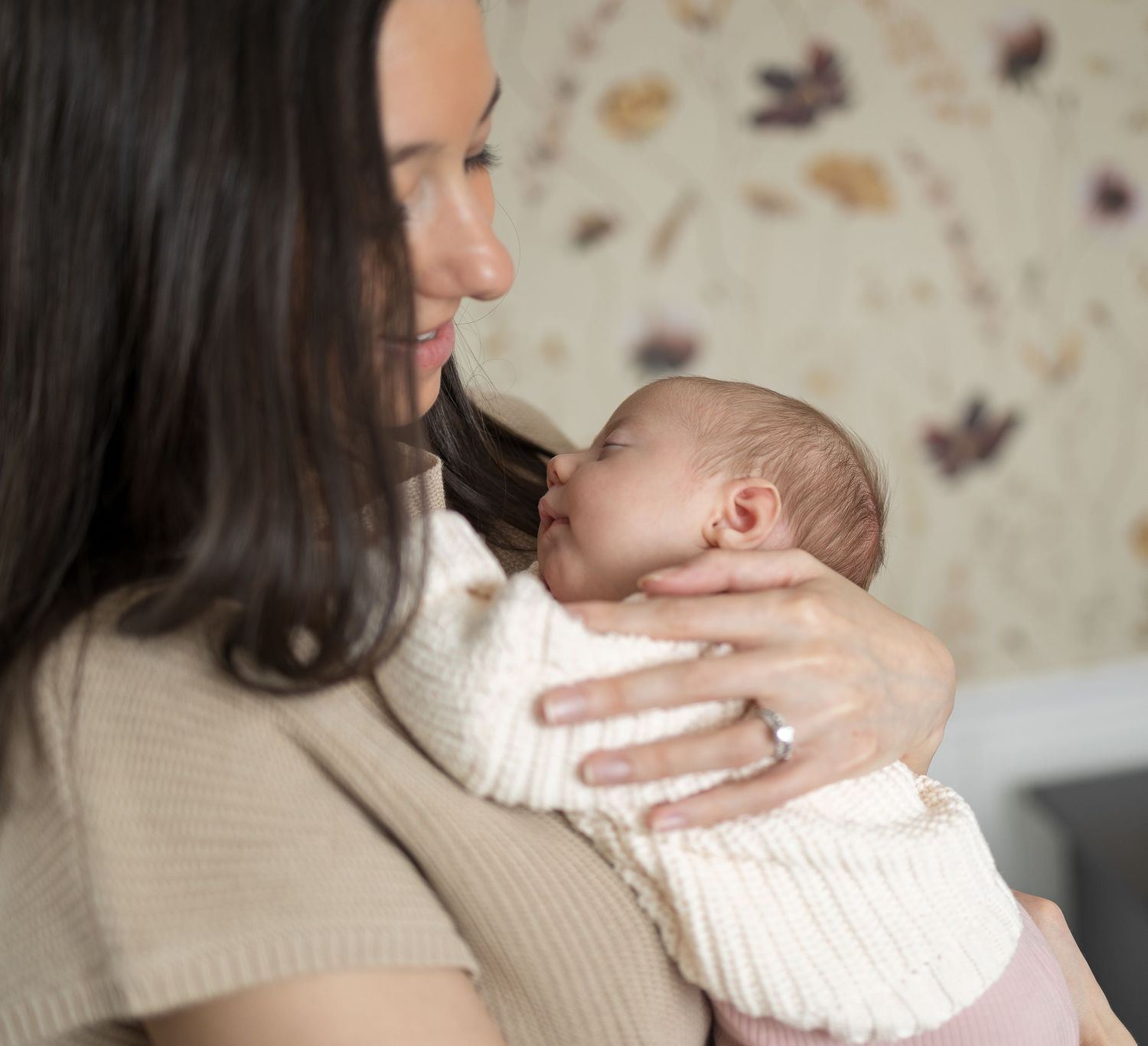Tender moment between parent and sleeping newborn against floral wallpaper.