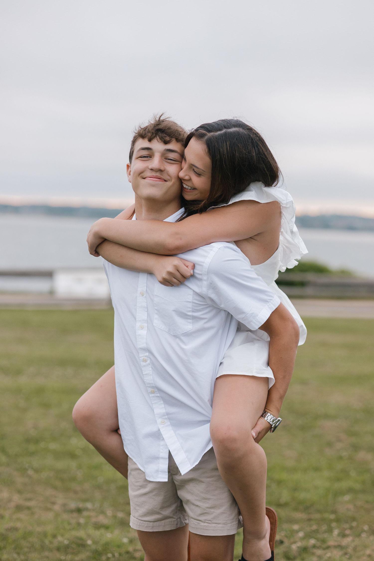 A playful couple in white outfits share joyful moments together on a grassy field by the water.