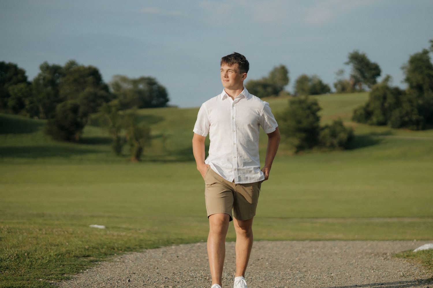 Person standing on path wearing white polo and khaki shorts with rolling hills in background.