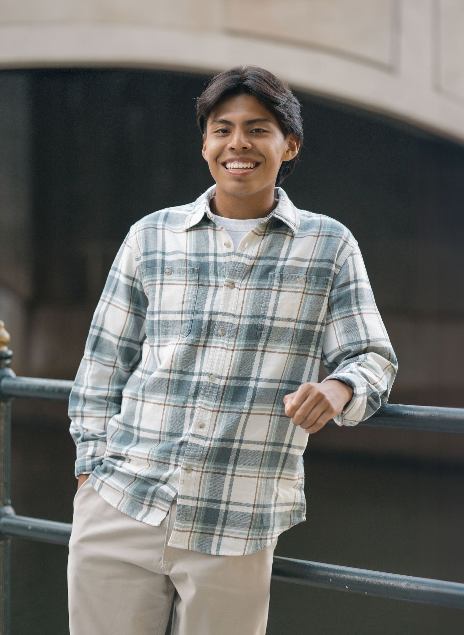 Person in plaid button-down shirt smiles while standing against railing outdoors.