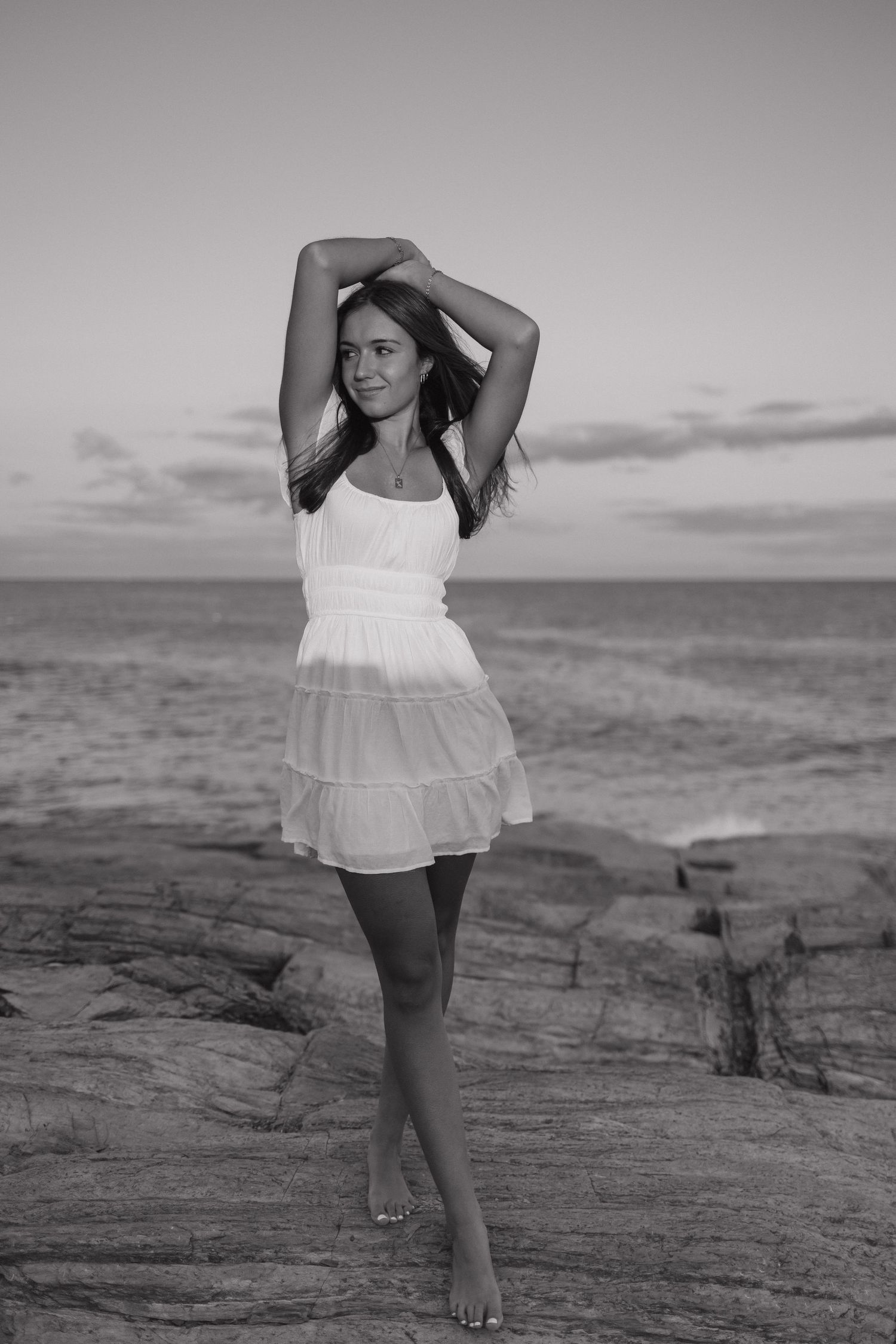 A flowing white dress creates an elegant silhouette against a coastal sunset in black and white.