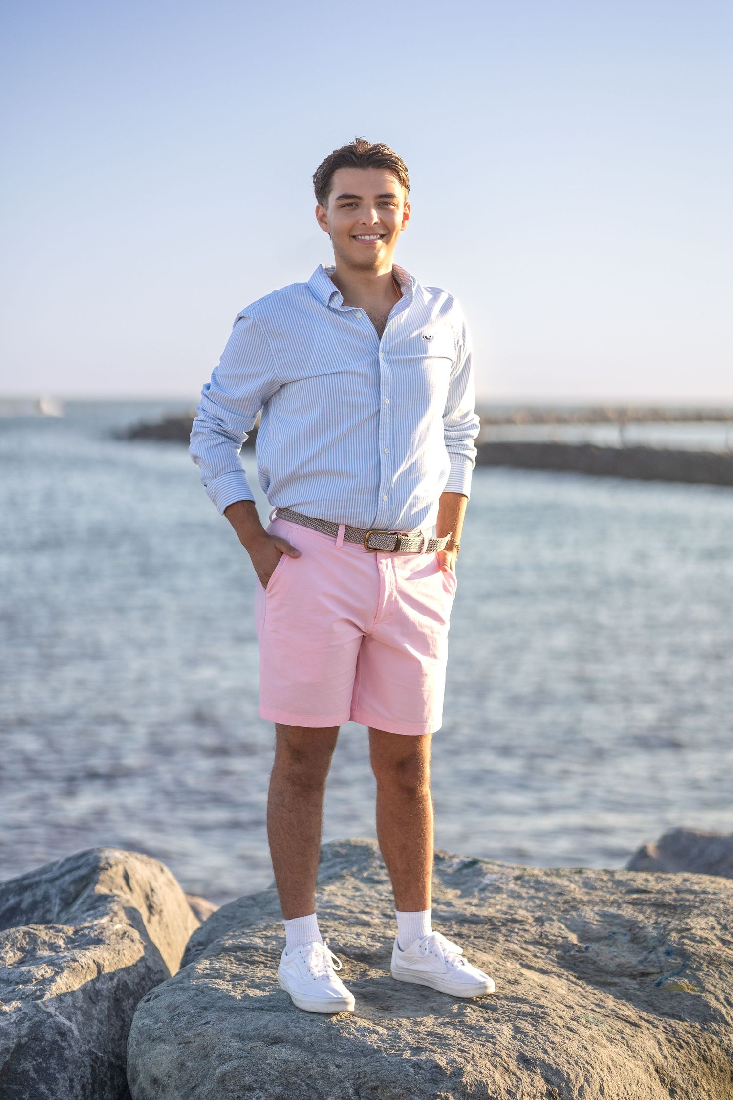 A person in a white button-down shirt and pink shorts stands on coastal rocks by the ocean wearing white sneakers.