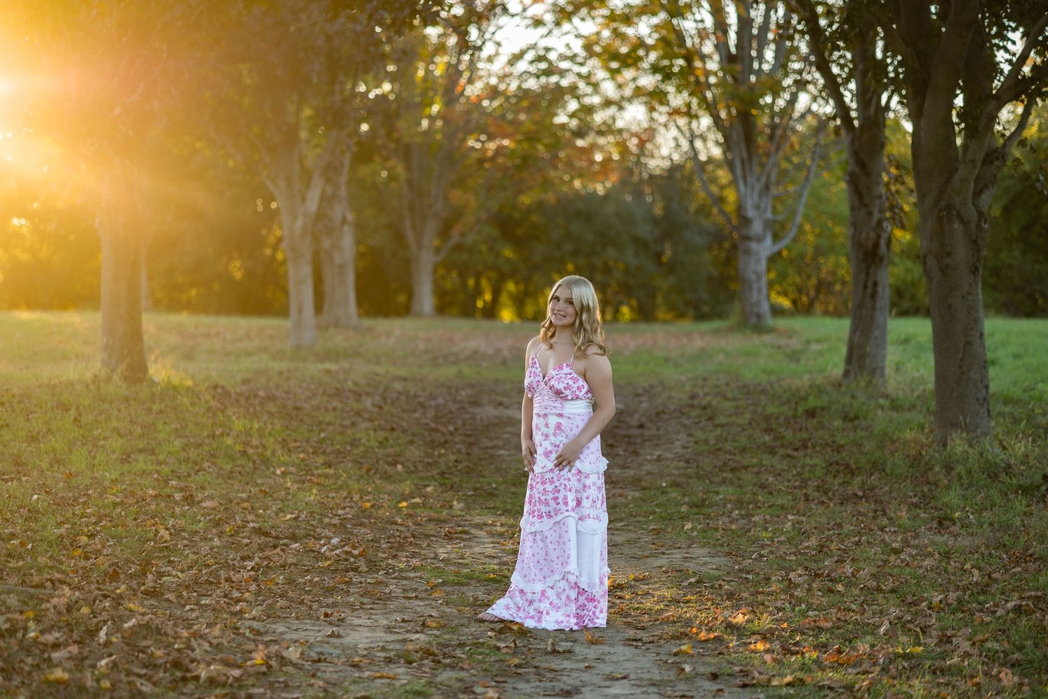 A person in a floral dress stands in a sunlit field during sunset with autumn trees in the distance.