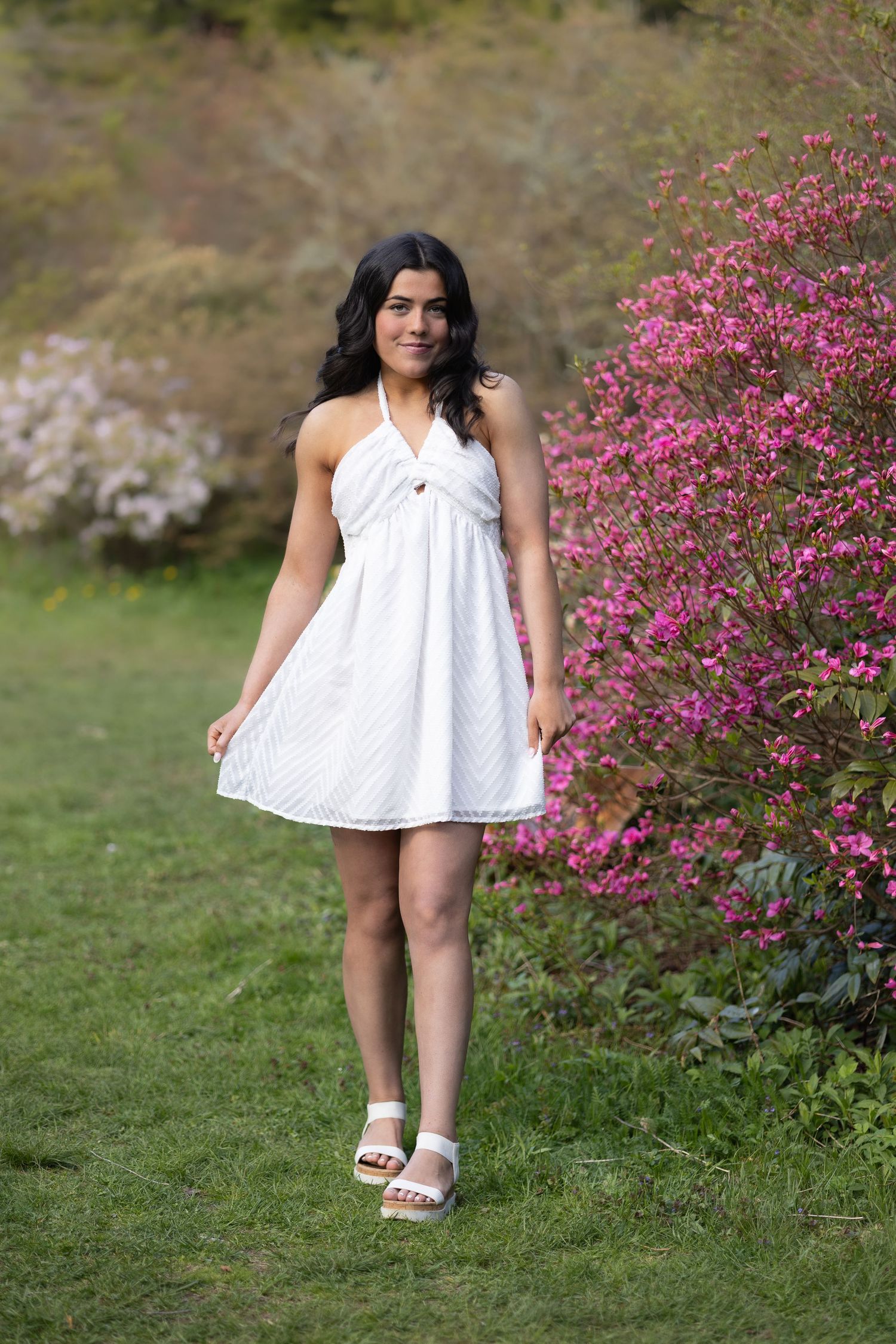 West Warwick senior photos in white halter dress and sandals standing near pink flowering bushes in a lush green garden in RI
