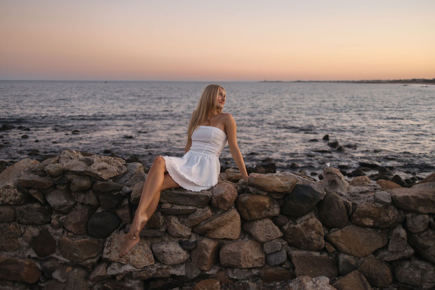 A person in a white dress poses on rocky oceanside wall during sunset.