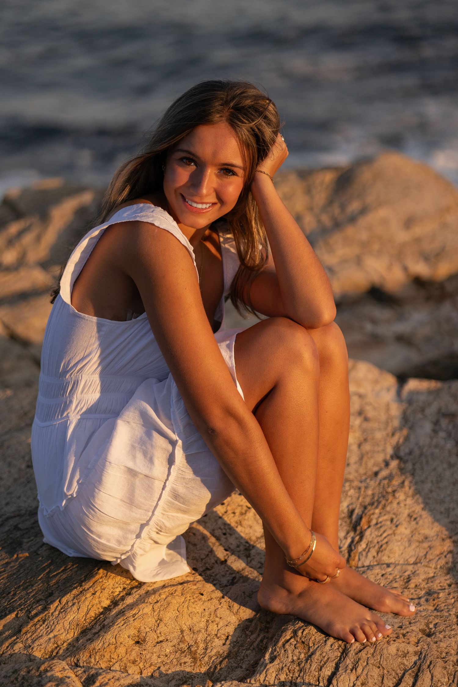 A white summer dress is worn while sitting on coastal rocks at golden hour.