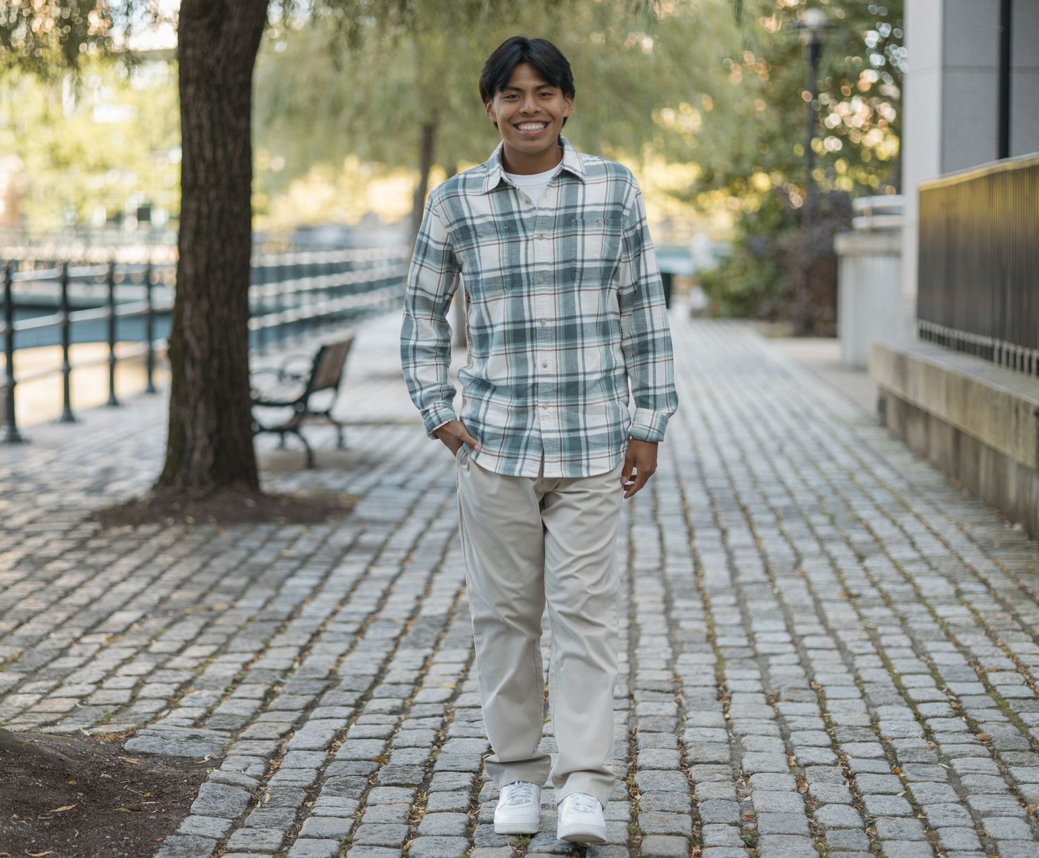 Person in plaid flannel shirt and khakis standing on brick pathway near trees.