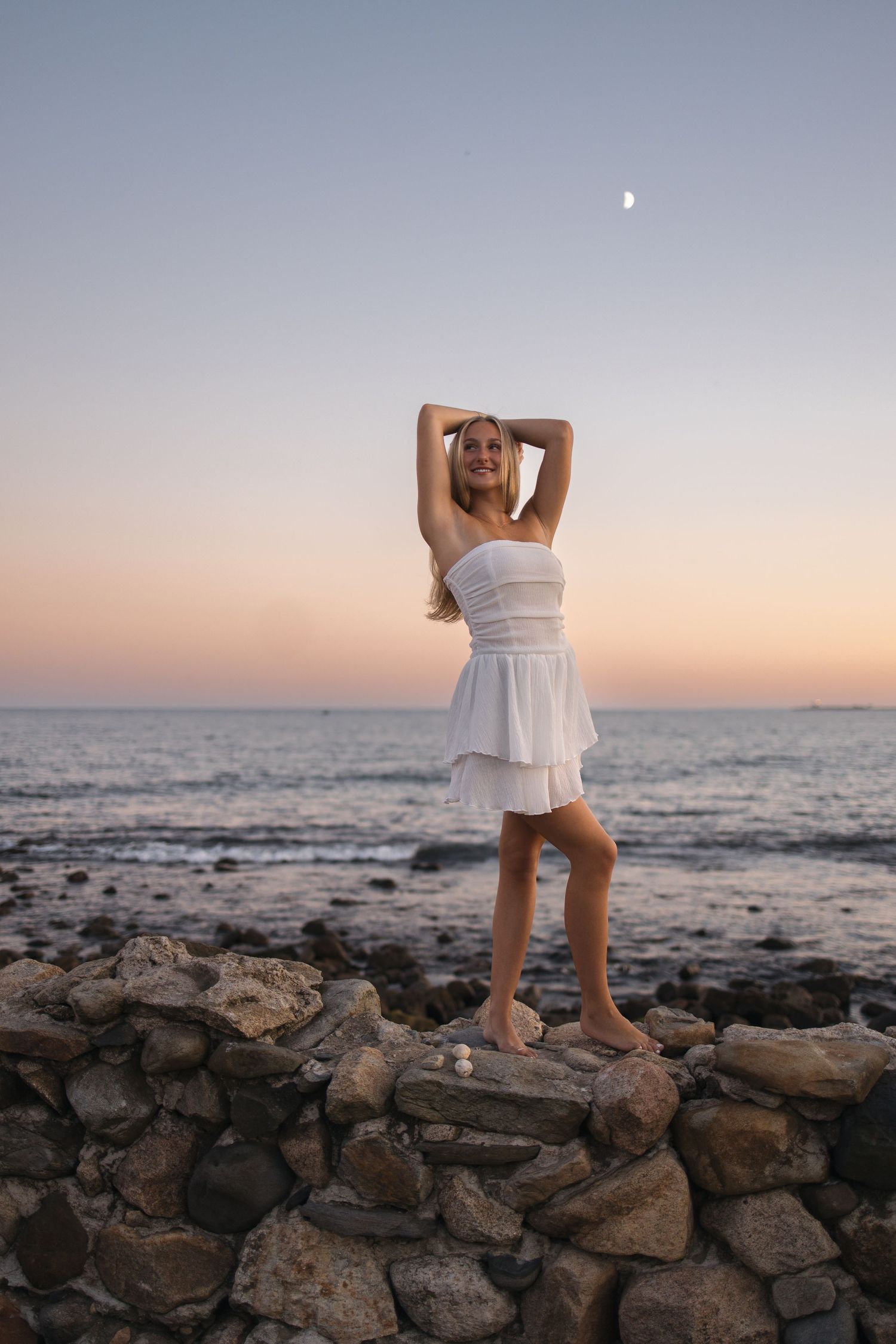 A figure in a white dress poses on a stone wall at sunset by the ocean with the moon visible in the sky.