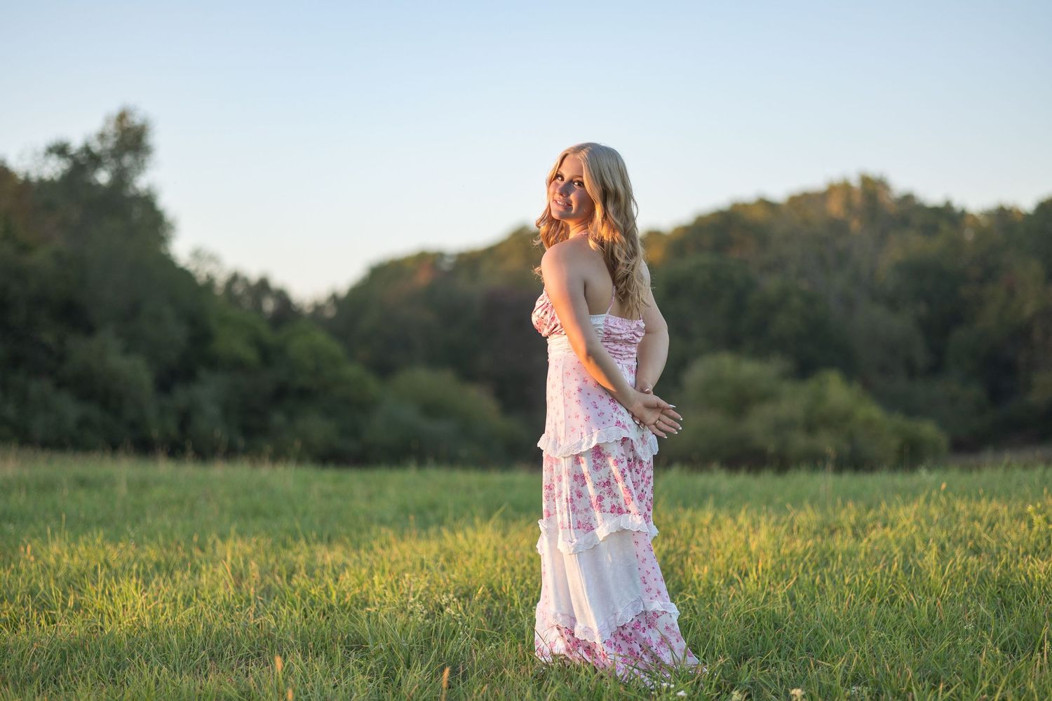 A woman in a flowing white dress stands gracefully in a sunlit meadow at golden hour.