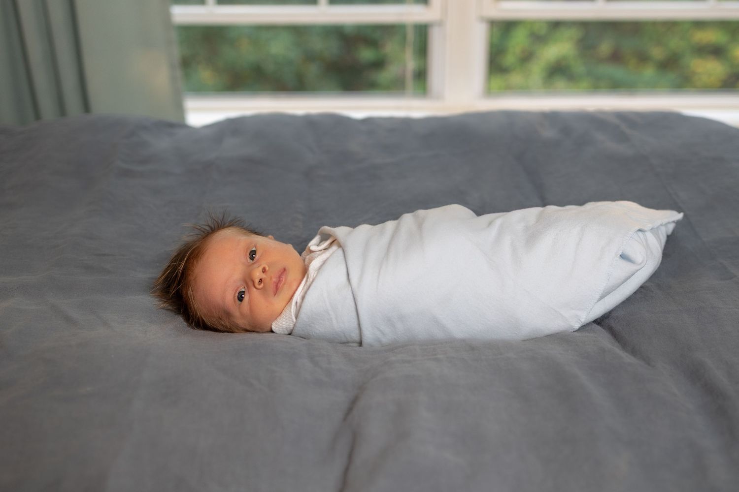 A baby in a white onesie lies on gray bedding while looking towards the side.