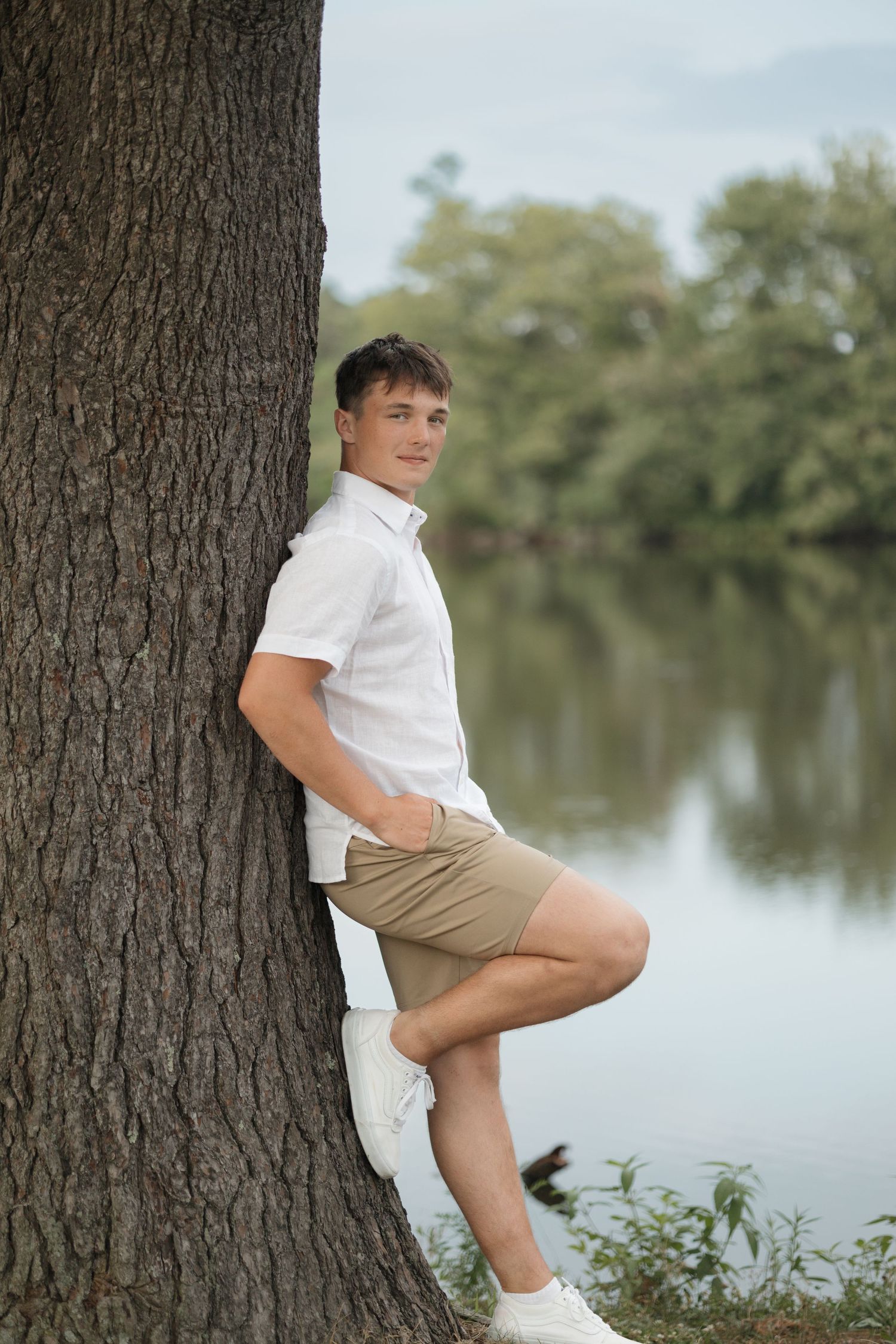 Someone leans against a tree trunk wearing white sneakers and casual summer clothing.