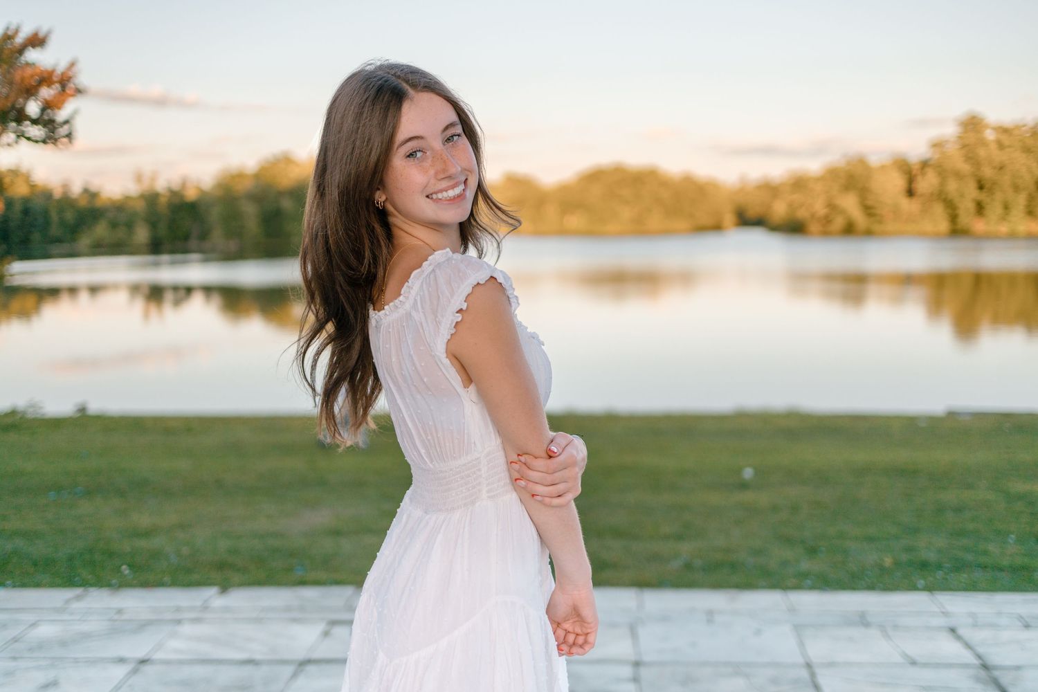 Woman in white summer dress spins joyfully by lakeside during sunset portrait session.