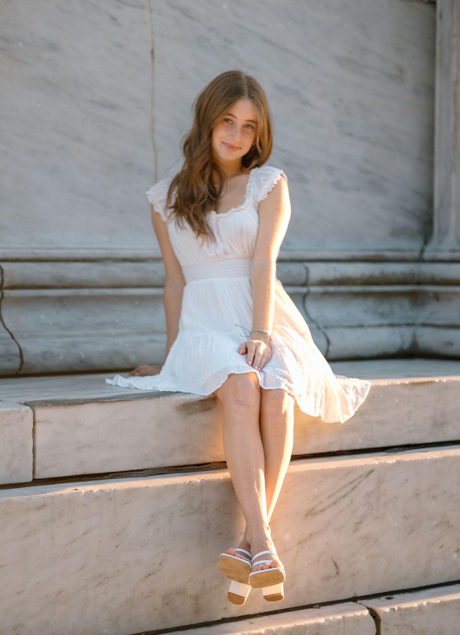A person in a white summer dress sits gracefully on marble steps in warm sunlight.