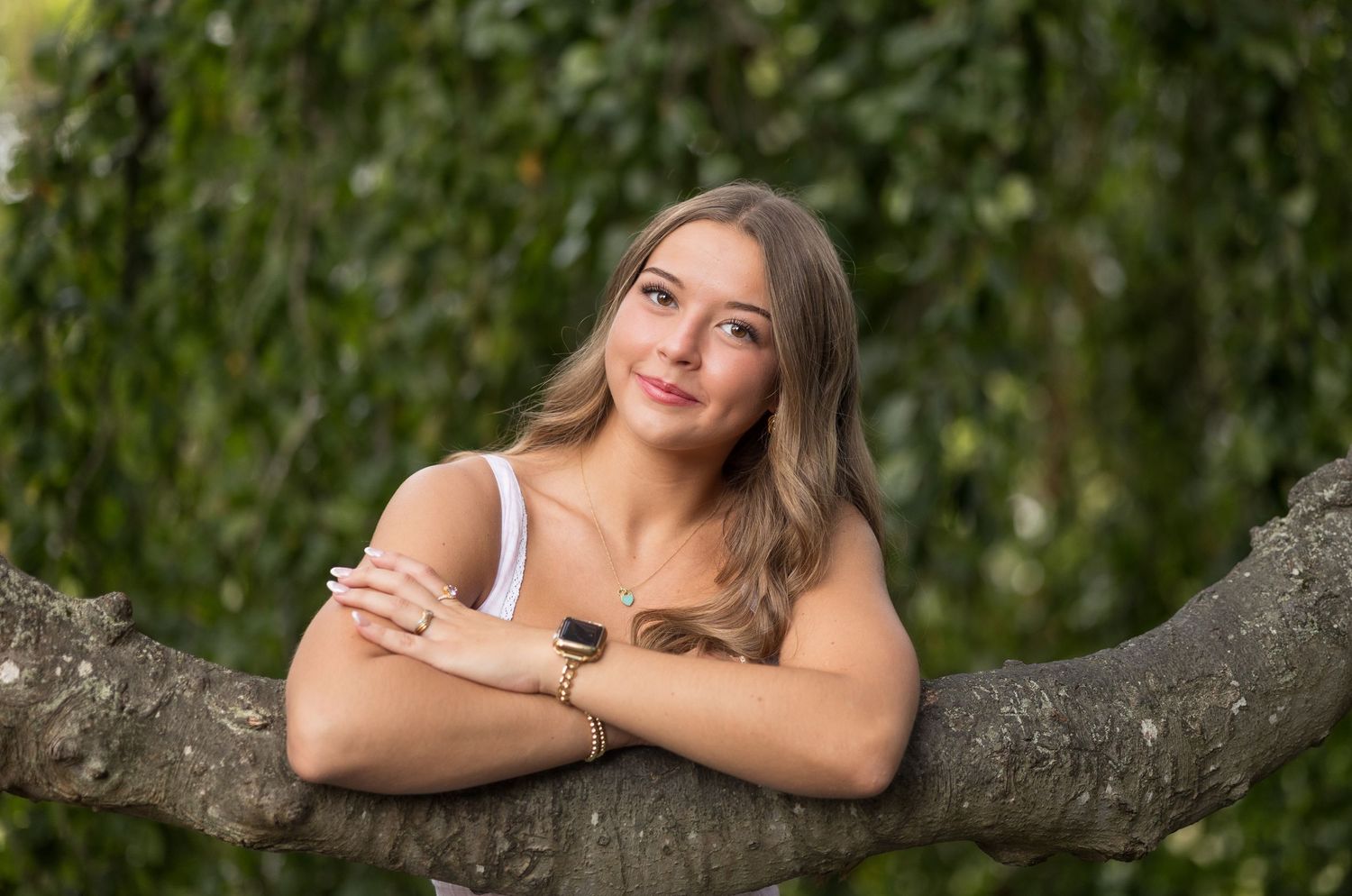 A woman in a white top poses gracefully on a tree branch in a lush green outdoor setting during a portrait session.