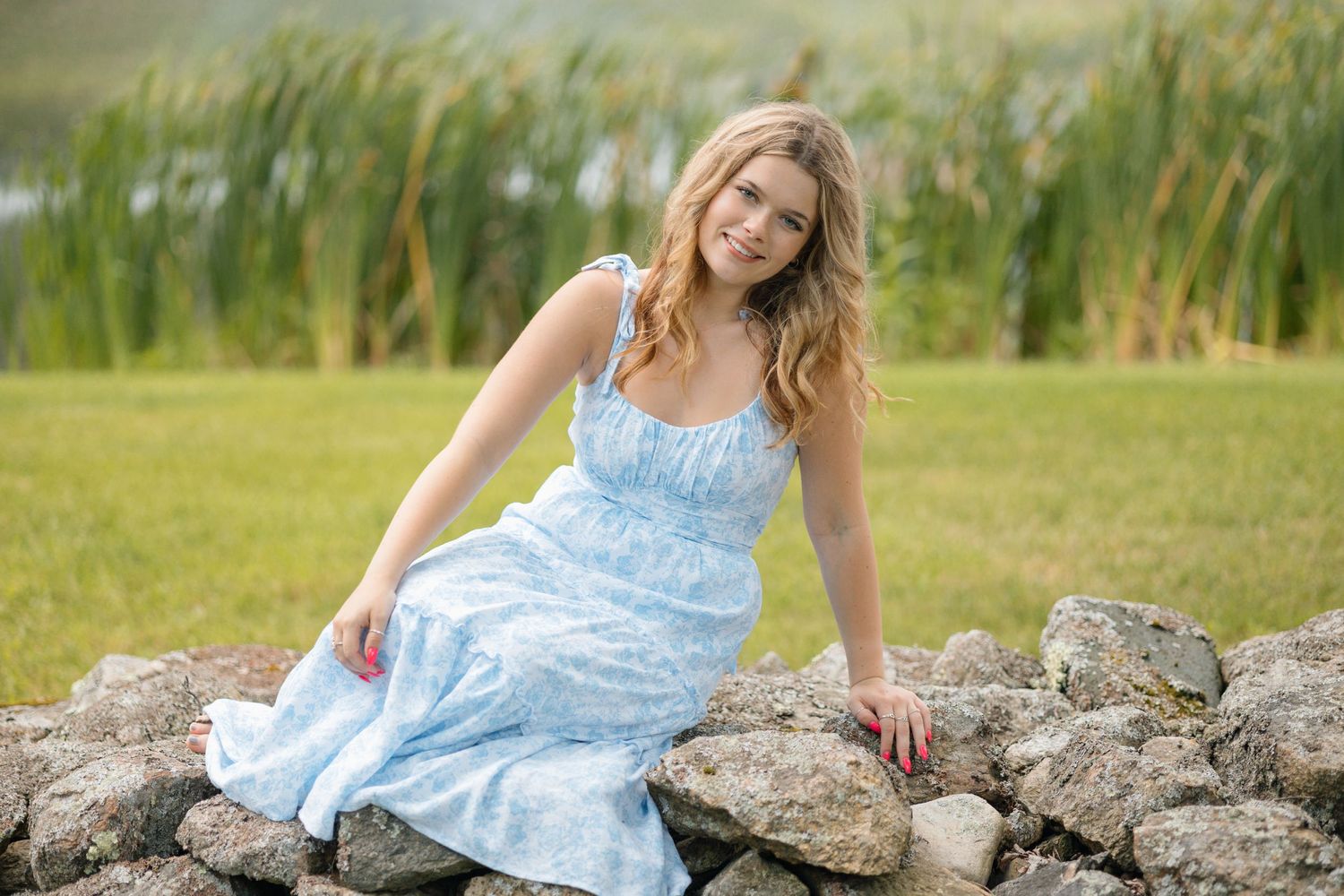 A person in a light blue dress sits casually on stone rocks near tall marsh grasses.