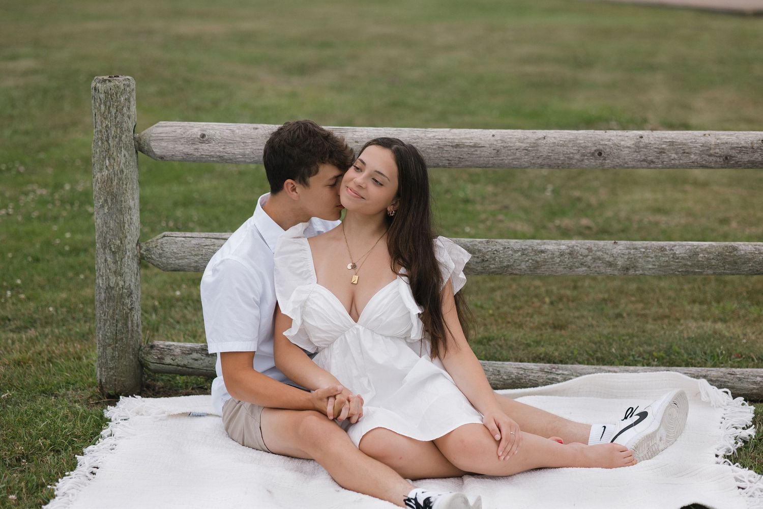 Romantic couple in white outfits sharing tender moments along a rustic wooden fence in a grassy field.