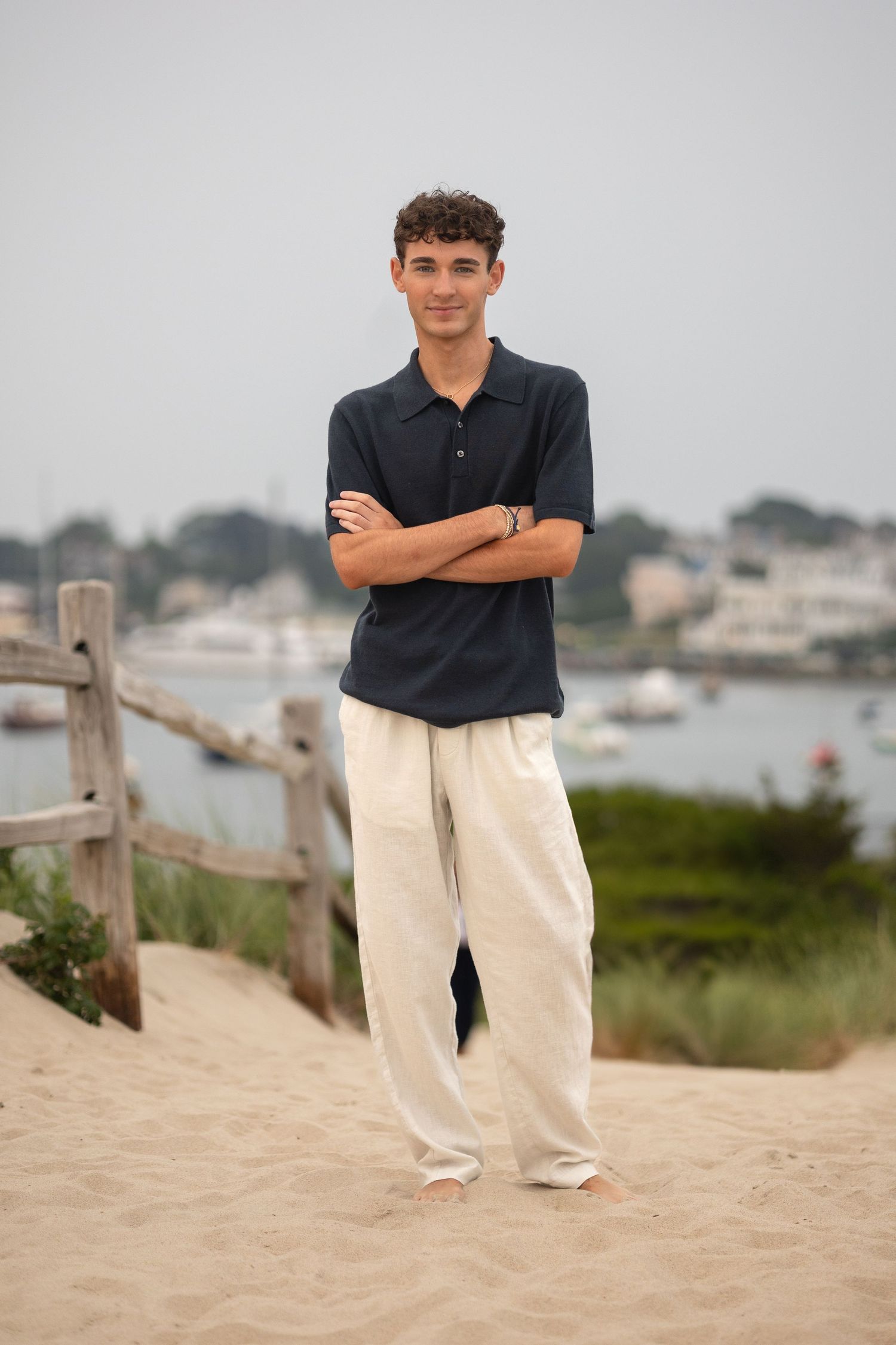 Model in navy polo and white pants poses on sandy beach with coastal scenery in background.