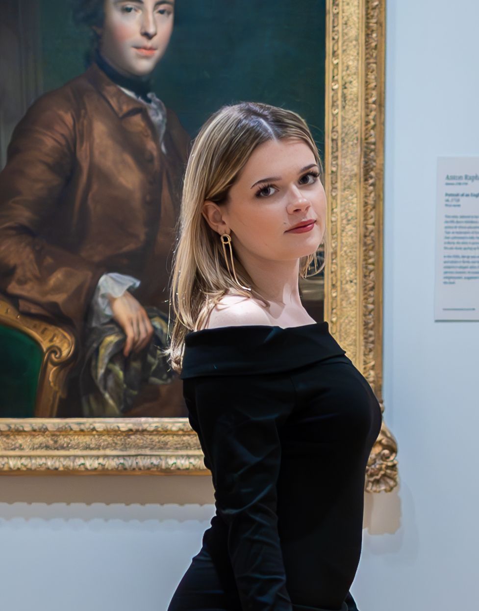 Figure in black dress poses next to classical portrait painting in ornate gold frame.