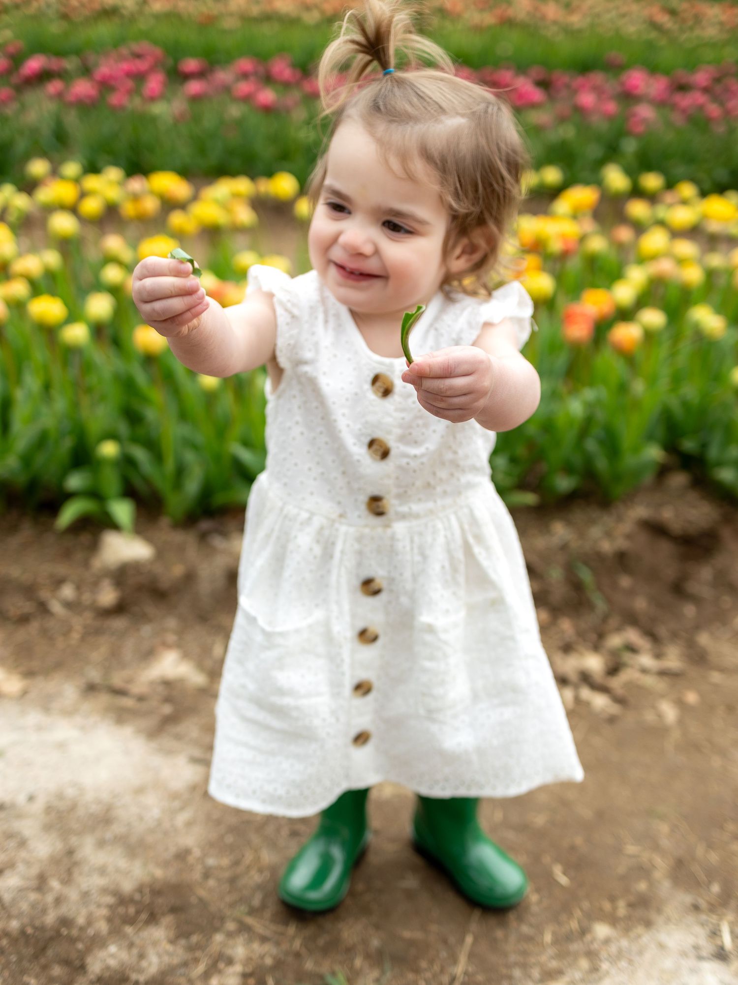 A toddler in a white dress and green boots smiles while playing in a flower garden.