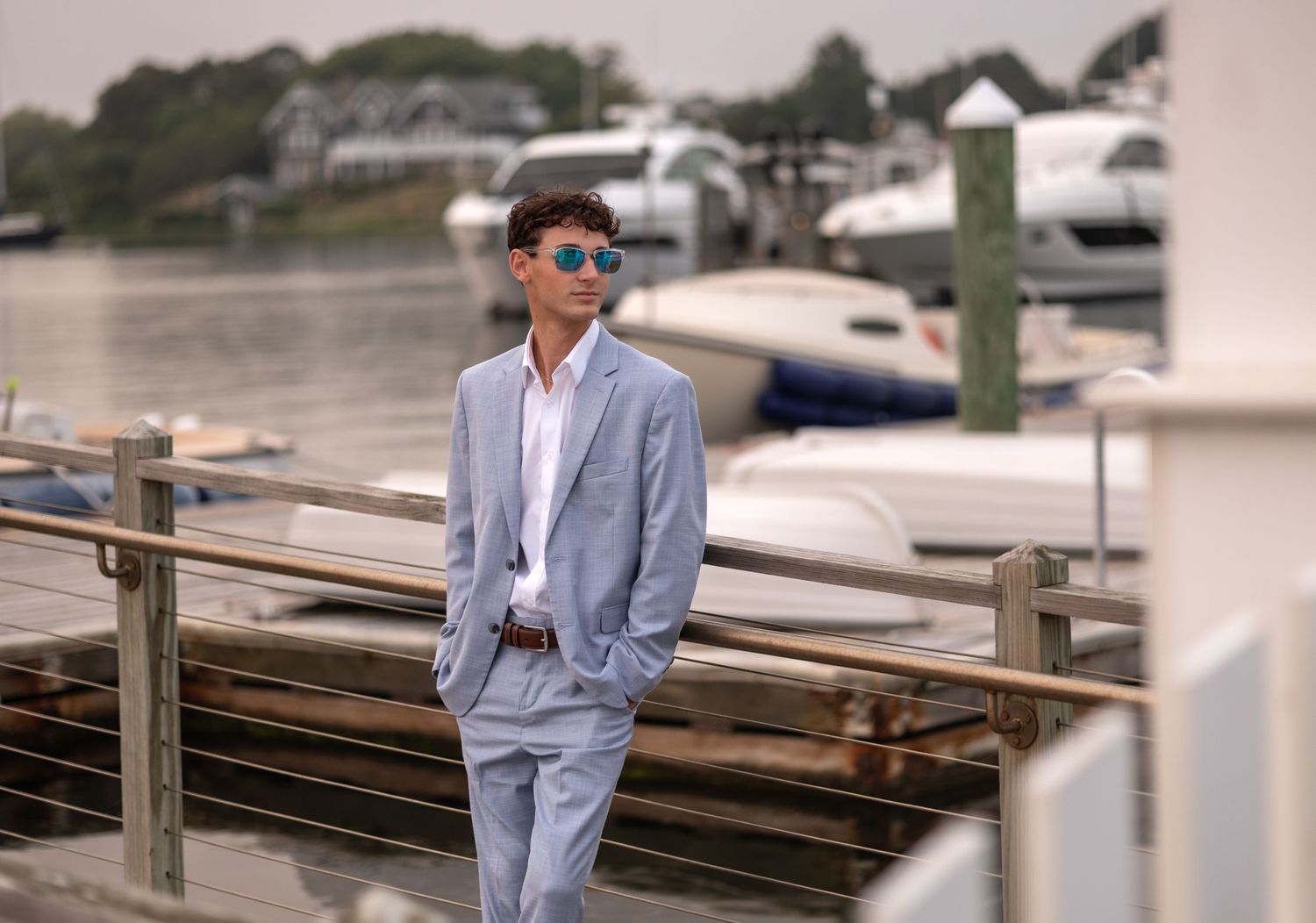 A high school senior in a light gray suit stands at a wooden dock near boats and water.