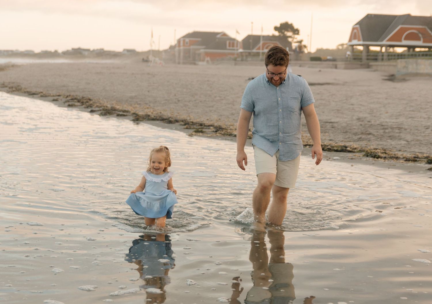 A parent and child splash together in shallow ocean water at sunset.