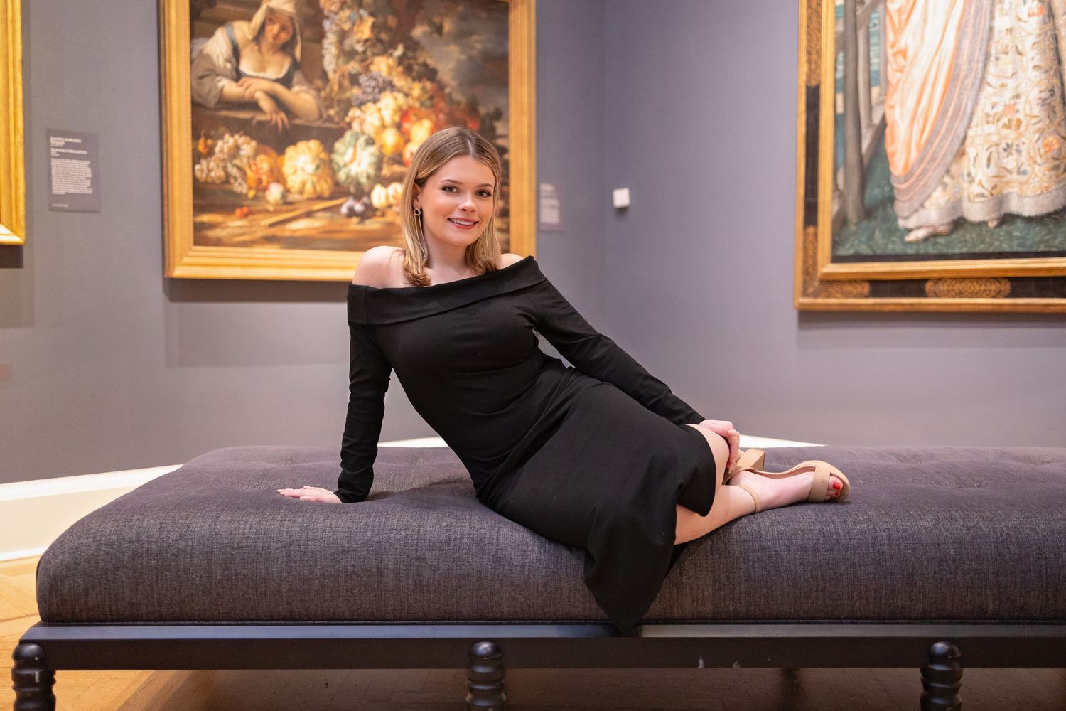 Woman reclines on dark gray bench in art gallery with classical paintings.