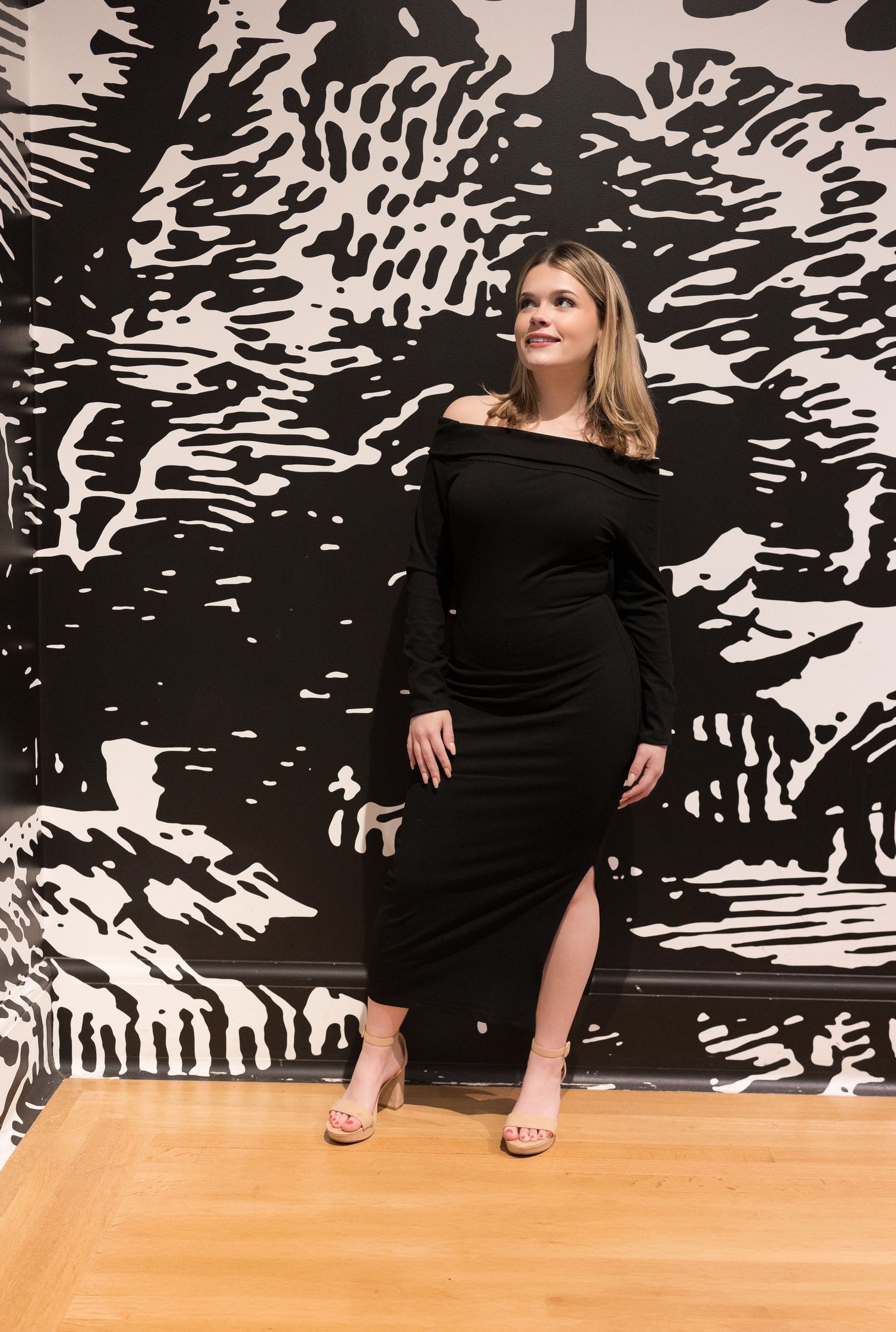 Elegant woman in black off-shoulder dress poses against large-scale monochromatic artwork.