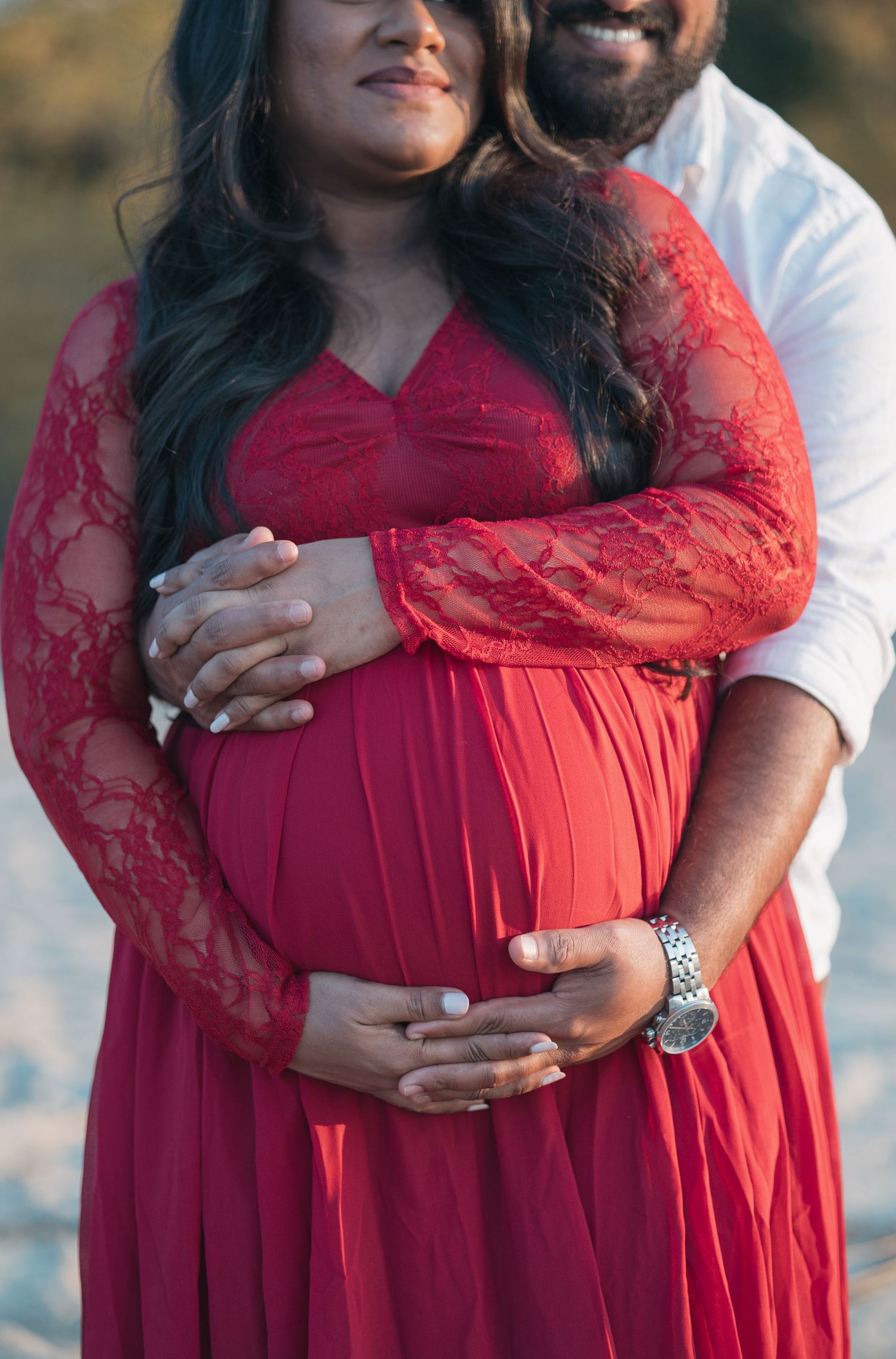 Close-up of couple's hands on pregnant belly in red lace maternity dress.