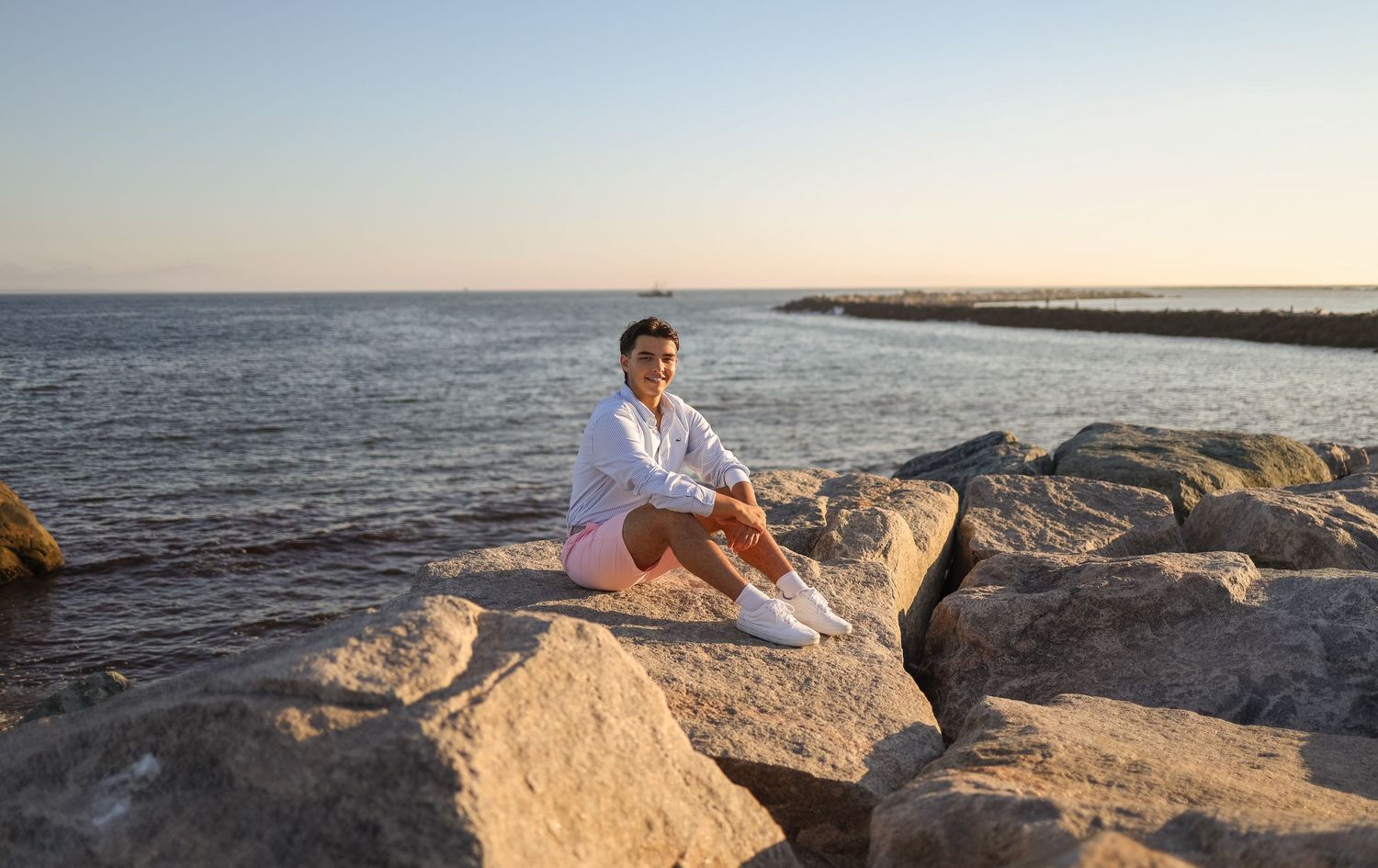 A person in white sits on coastal rocks at sunset by the ocean, creating a peaceful silhouette against the sky.