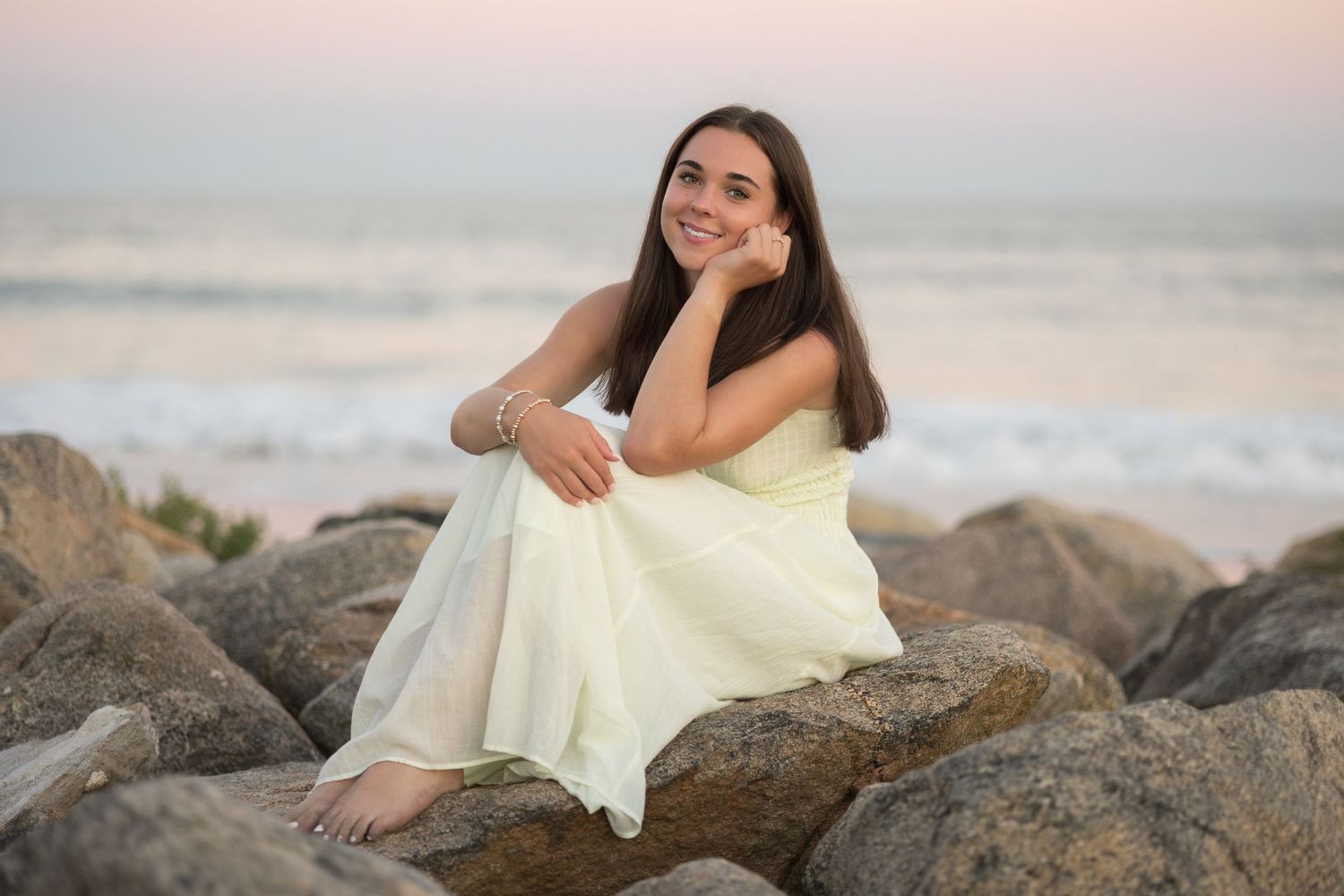 Woman in flowing white dress poses gracefully on rocky beach during sunset photoshoot.