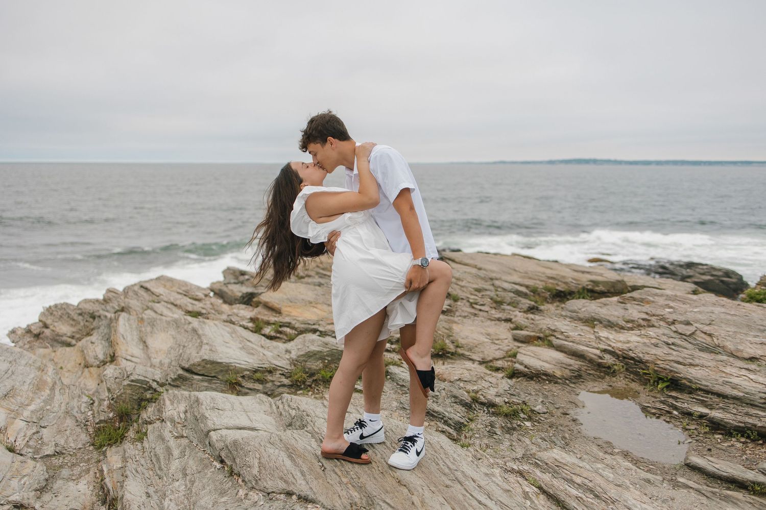 Couple in white clothing embracing on rocky coastal cliffs with ocean waves in the background.