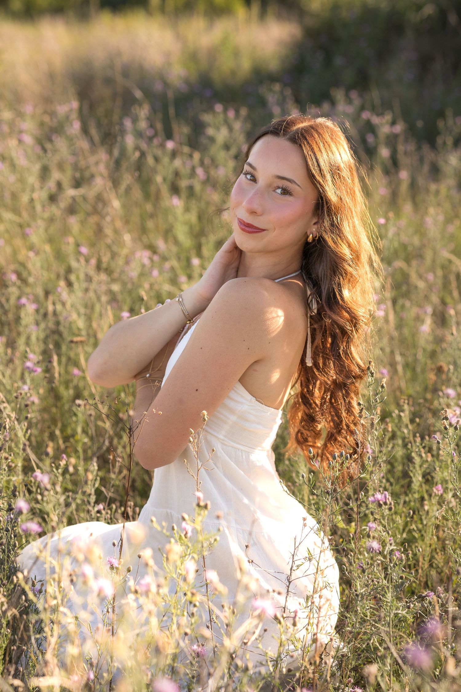 A person in a white dress poses gracefully among wildflowers in a sunlit meadow during golden hour.