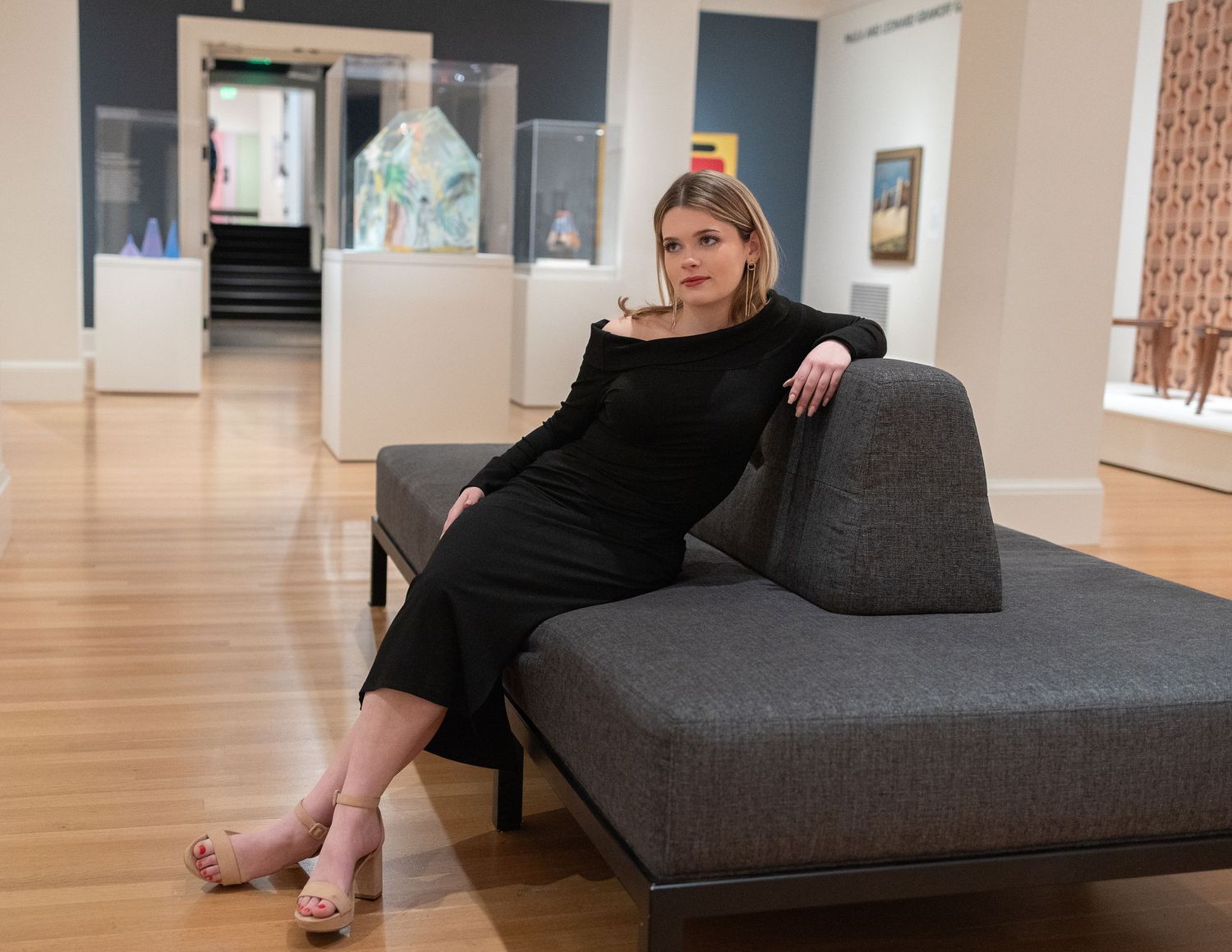 Woman in black dress lounges on grey modern sofa in art gallery setting.