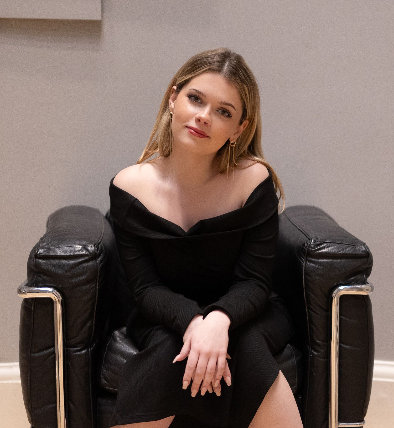 Person in black off-shoulder dress poses in leather armchair against neutral wall.