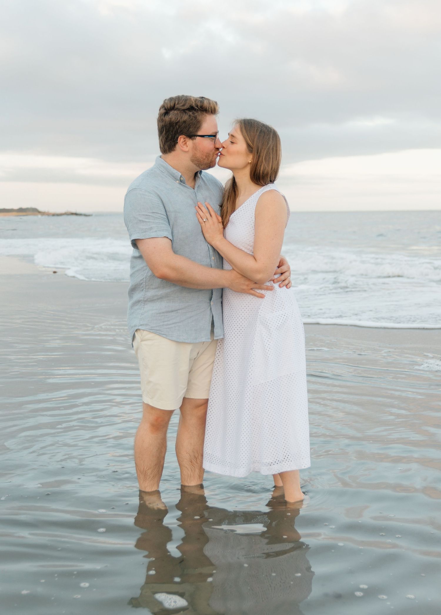 A couple shares a romantic kiss while standing in shallow ocean water at sunset.