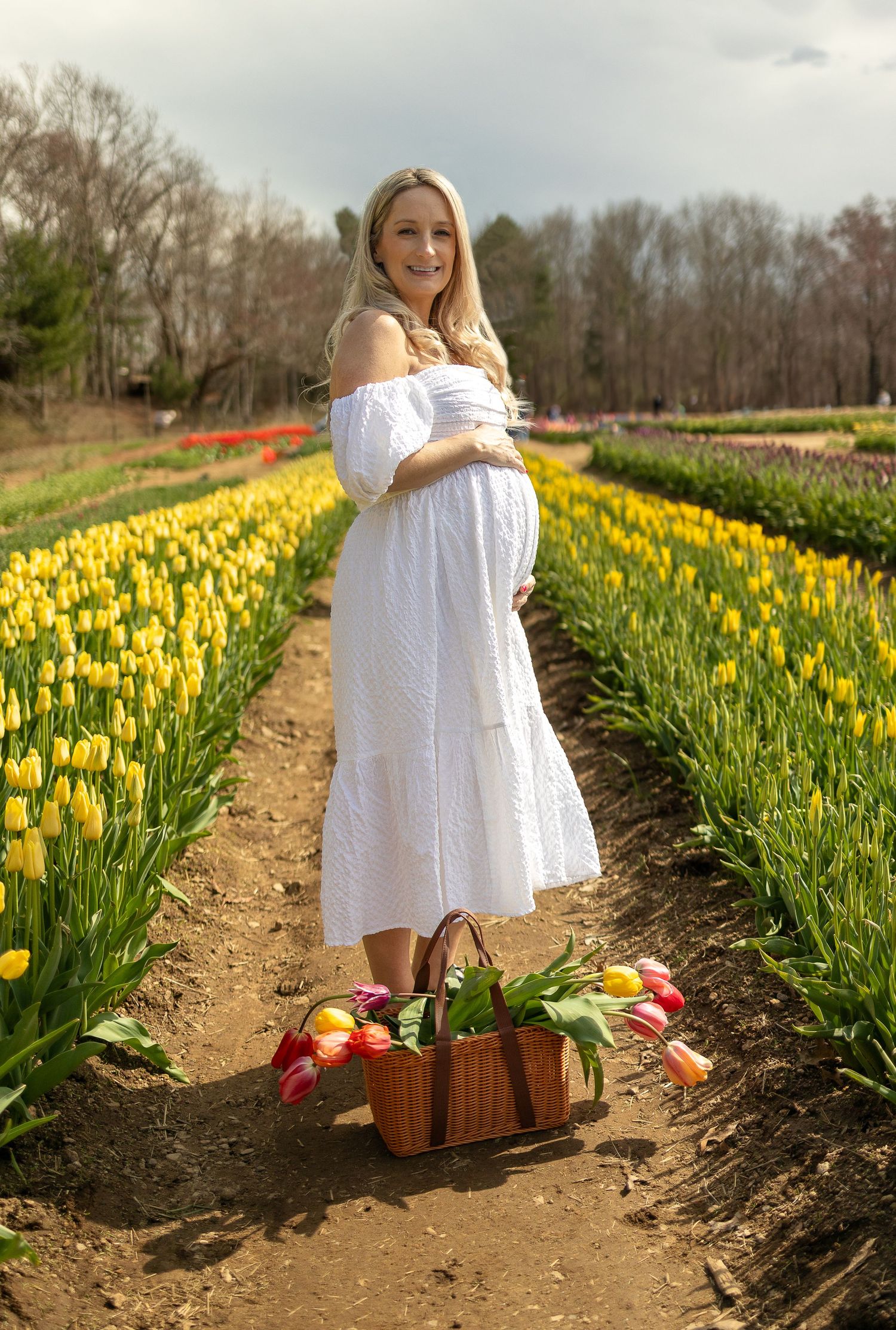 Expectant mother in white dress standing in tulip field with basket of fresh flowers.