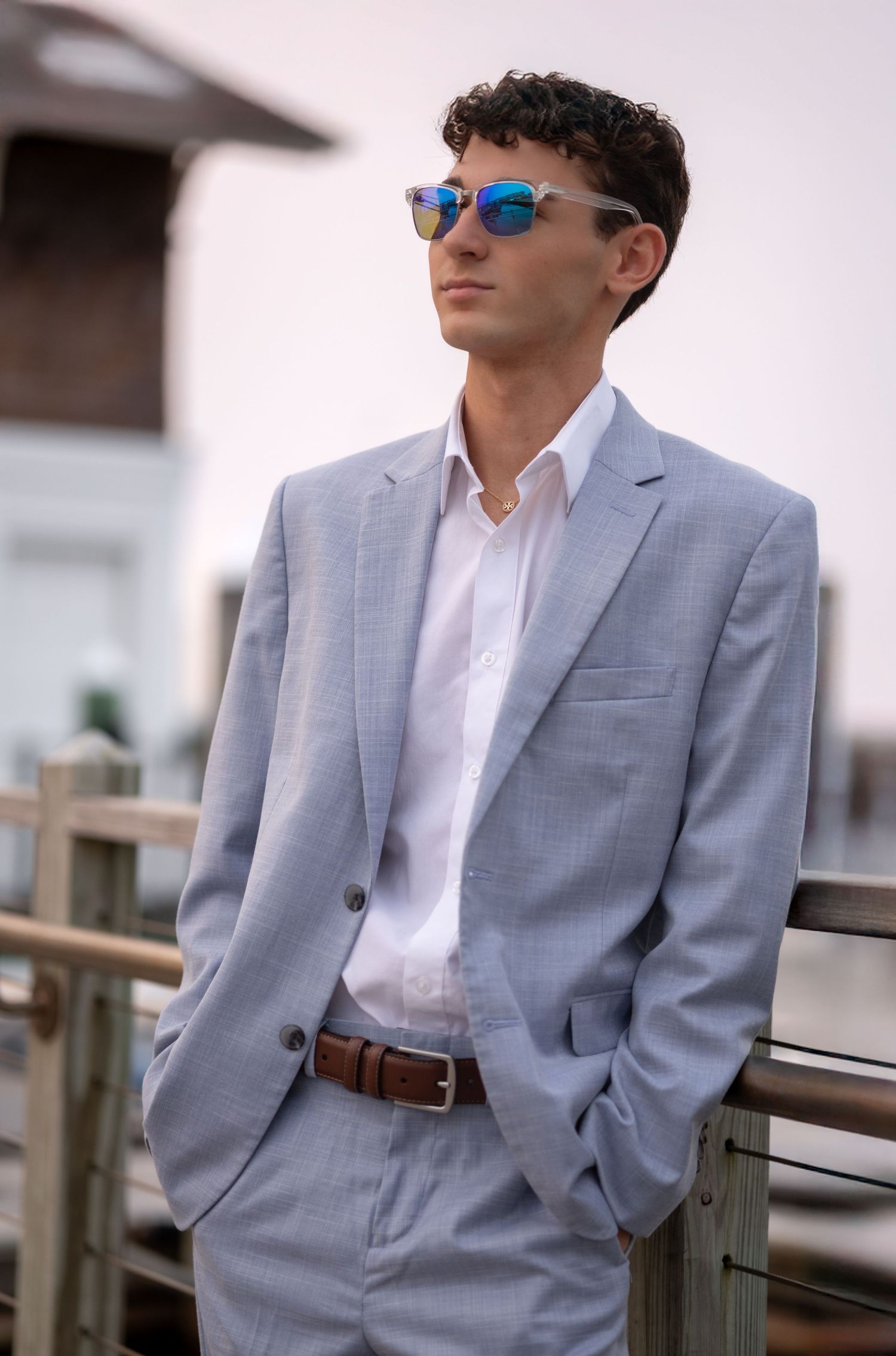 Model wearing light gray suit jacket and sunglasses poses against white railing in outdoor setting.