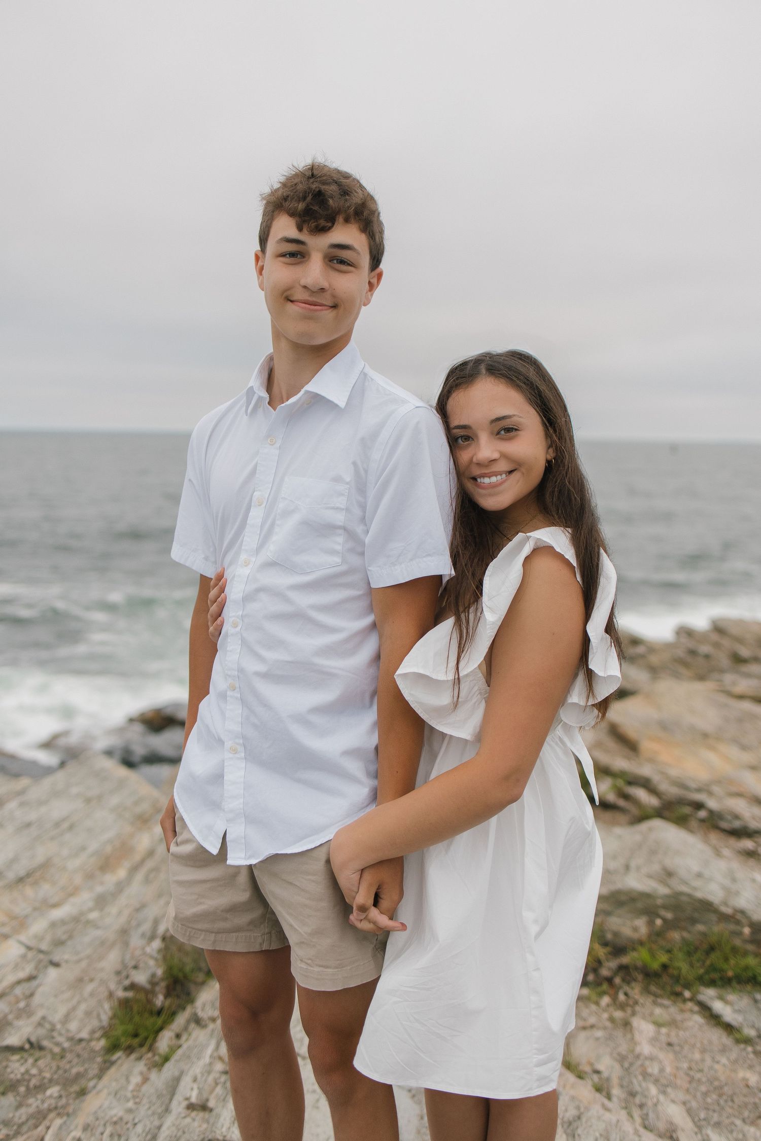 Couple dressed in white embracing on rocky oceanside cliffs with waves and cloudy sky.
