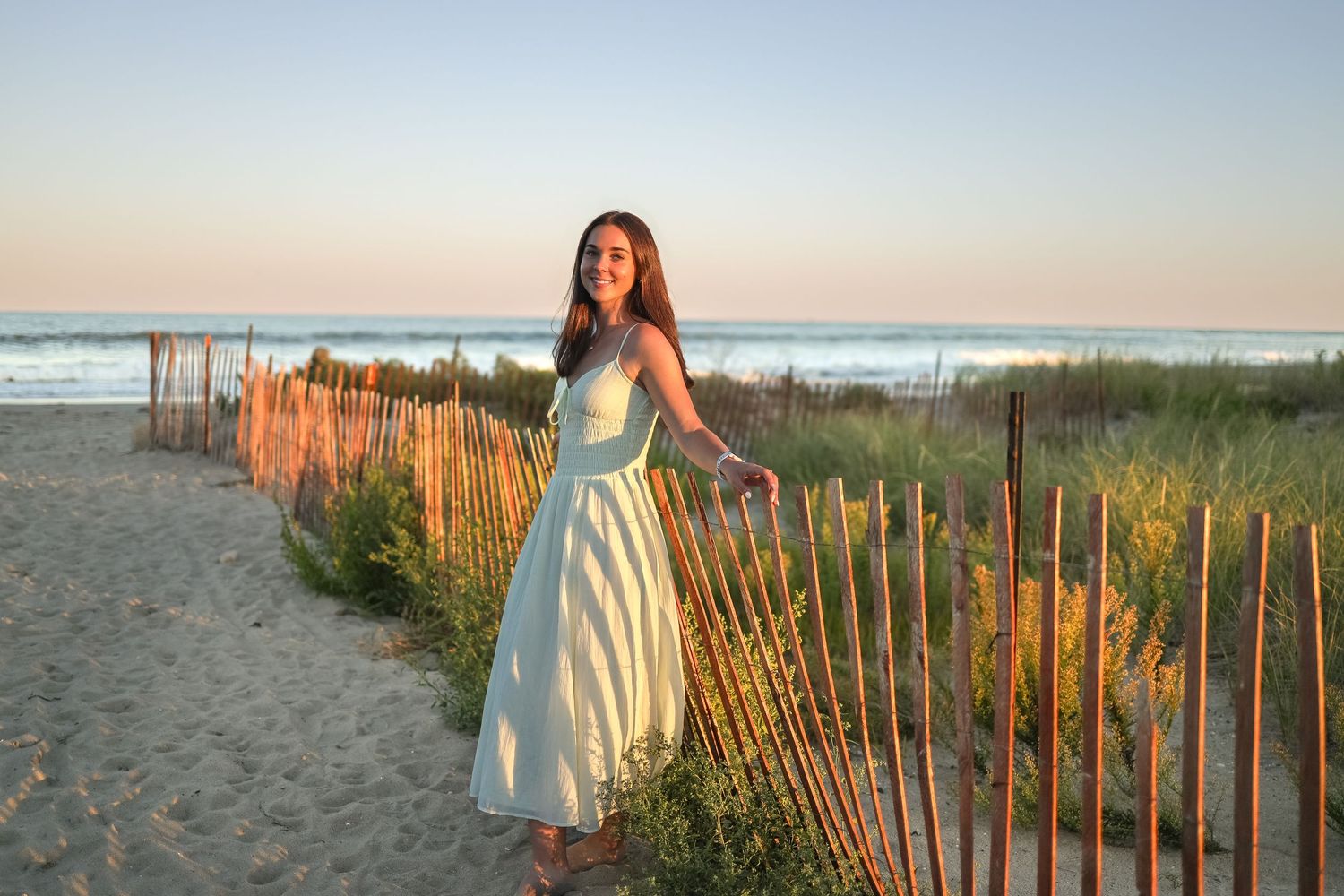 A person in a yellow sundress stands by wooden beach fences at sunset along a sandy coastal shoreline.