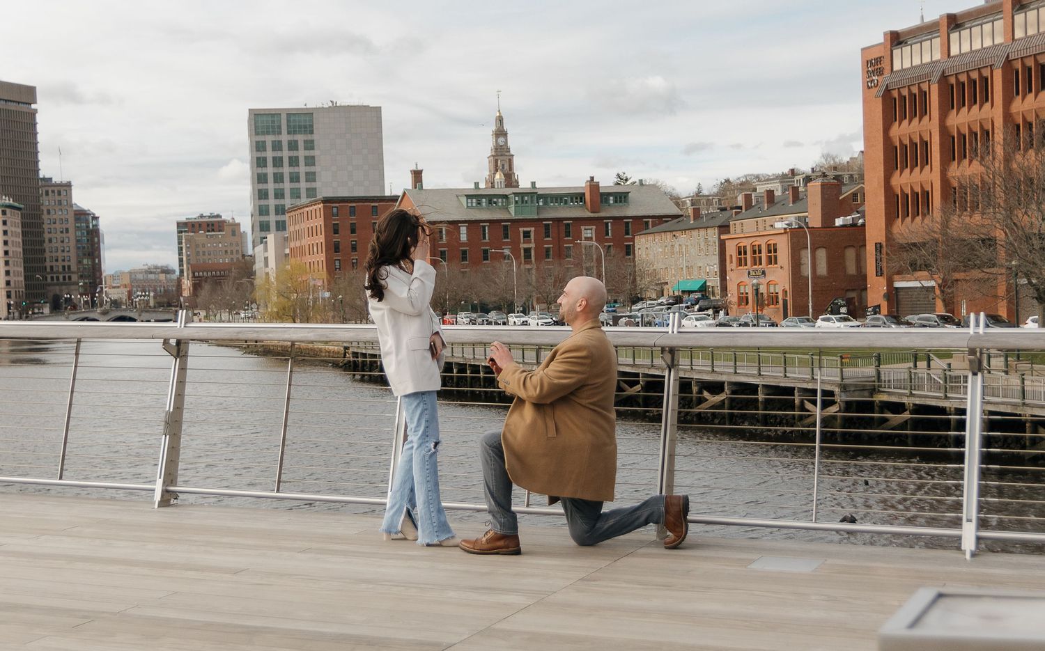 A proposal moment on a waterfront boardwalk with historic brick buildings in the background.