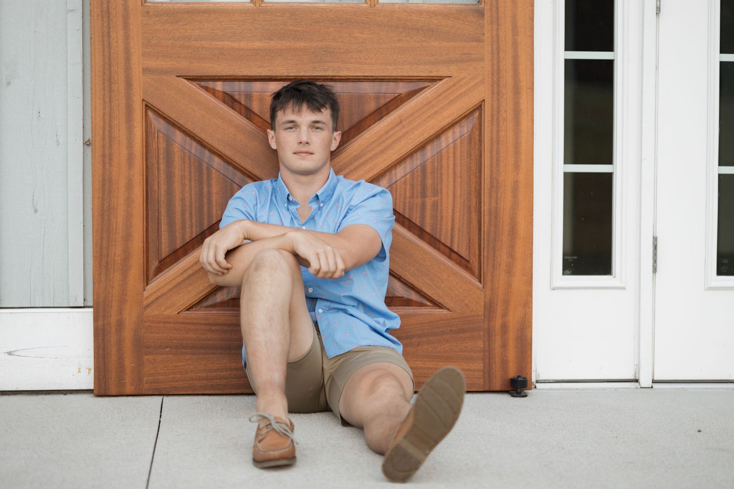 Person sitting on ground against wooden door wearing light blue shirt and brown shorts.