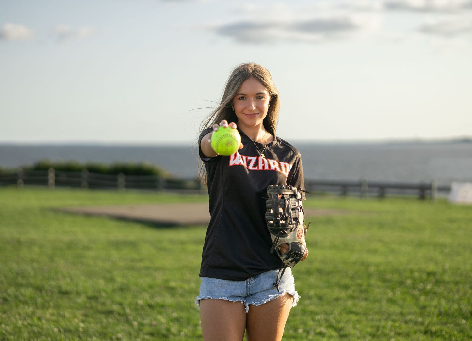 A softball player in a dark t-shirt holds up a yellow ball while standing in a grassy field.