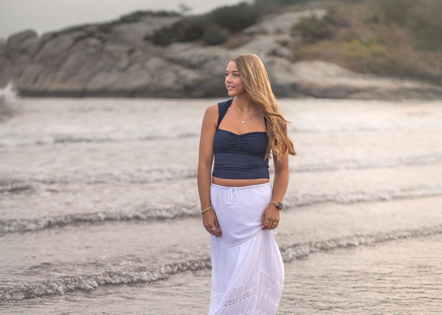 A person in a navy crop top and white maxi skirt walks along a rocky beach at sunset.